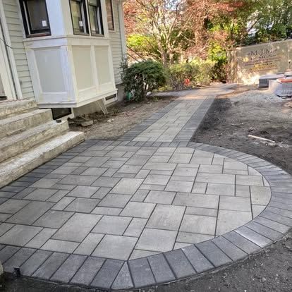 Curved gray paver walkway beside a house leading to a stone wall and garden.