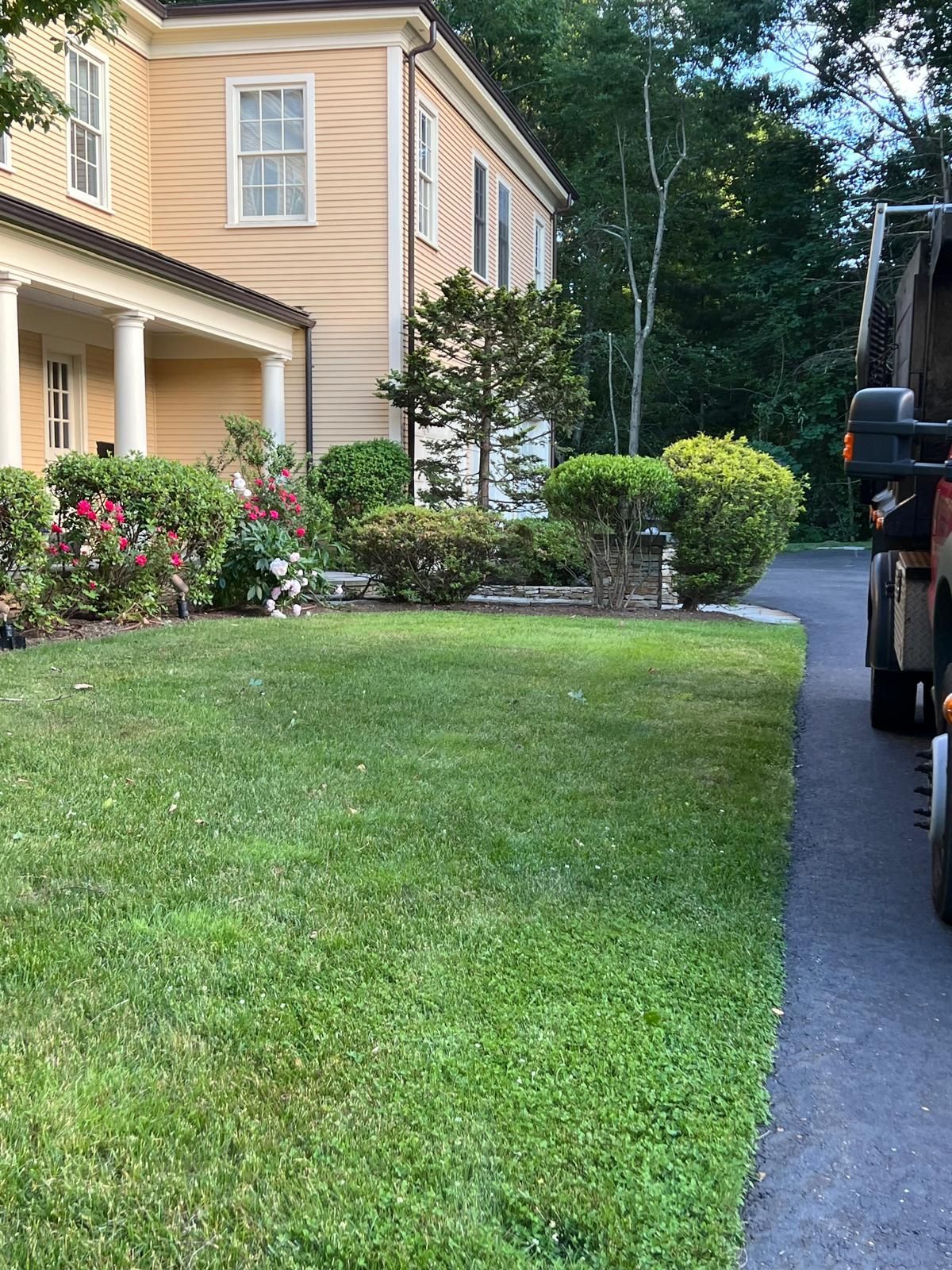 Front yard with green lawn, beige house, shrubs, and a driveway with a parked vehicle.
