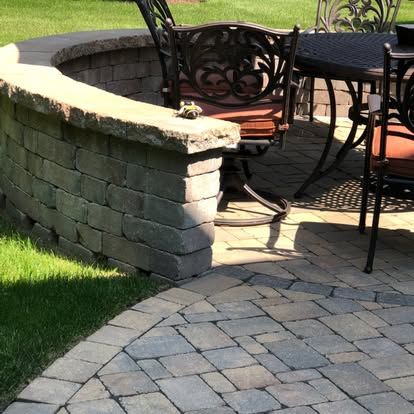 Curved stone patio wall with outdoor chairs and table beside a paved walkway and lawn