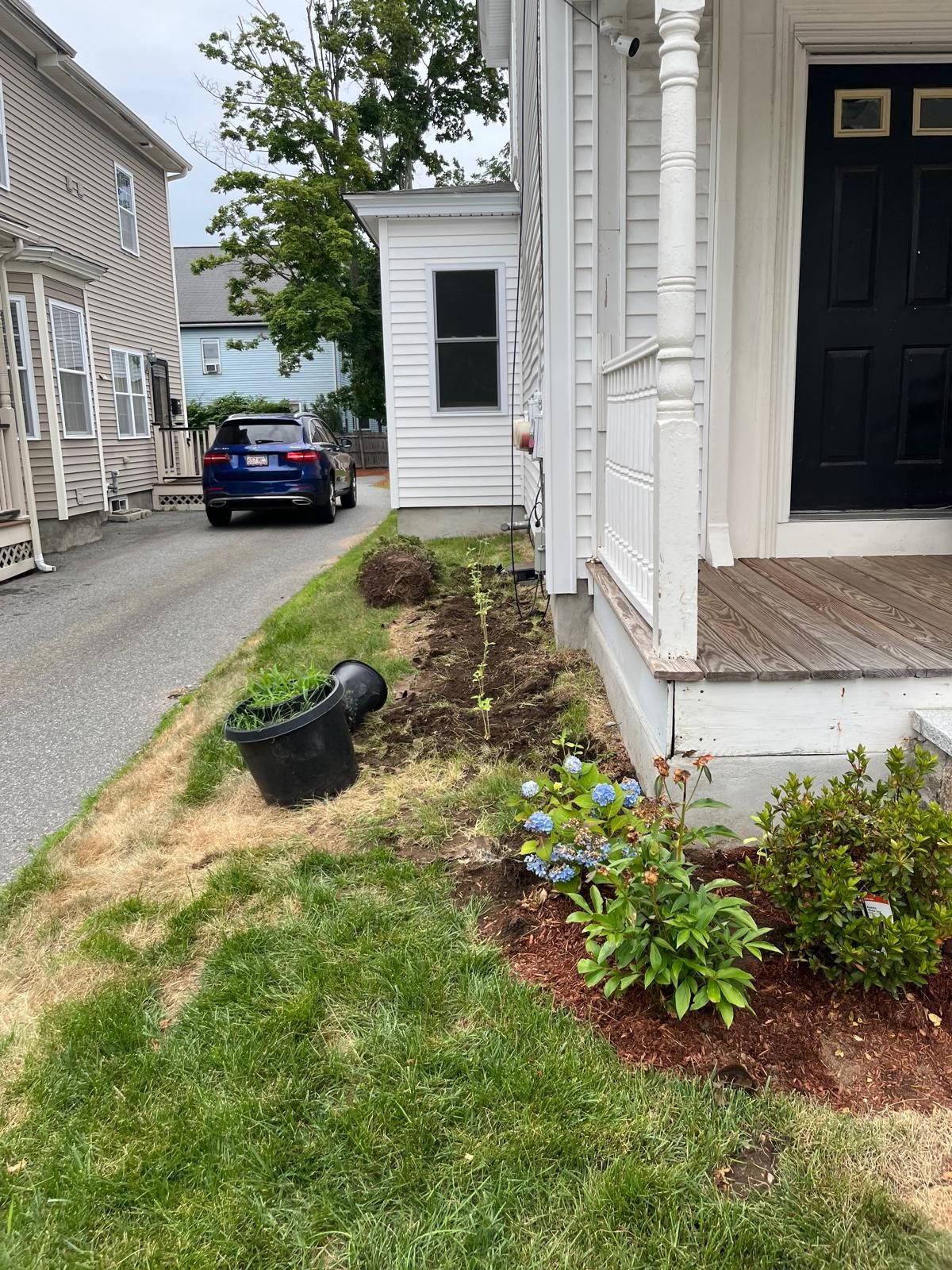 Front porch and garden bed beside a white house, with a blue car parked on a narrow street.