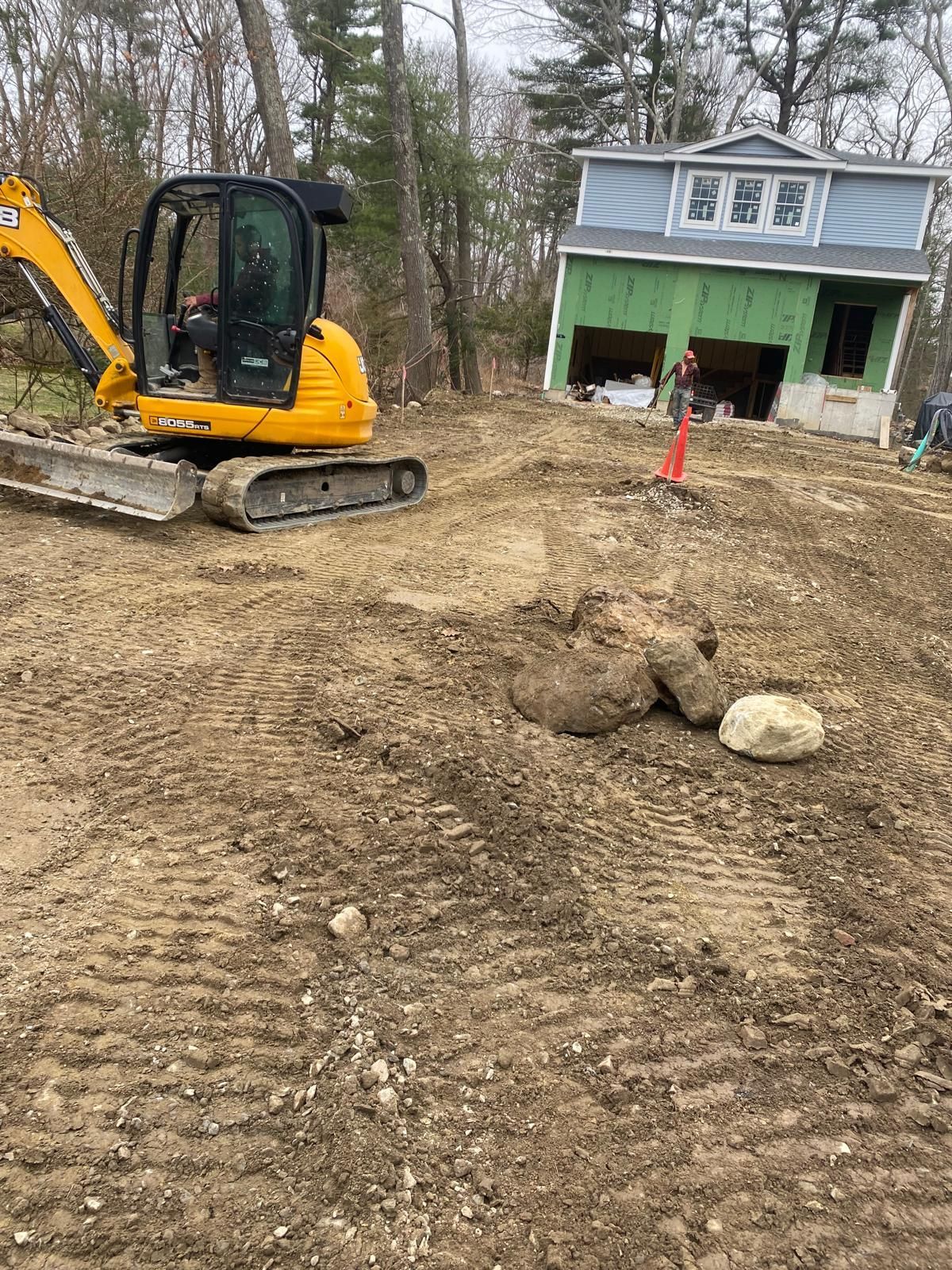 Yellow excavator leveling dirt in a yard beside a green house under construction.