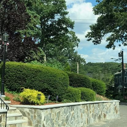 Stone retaining wall beside manicured hedges and trees along a quiet roadside under a cloudy sky