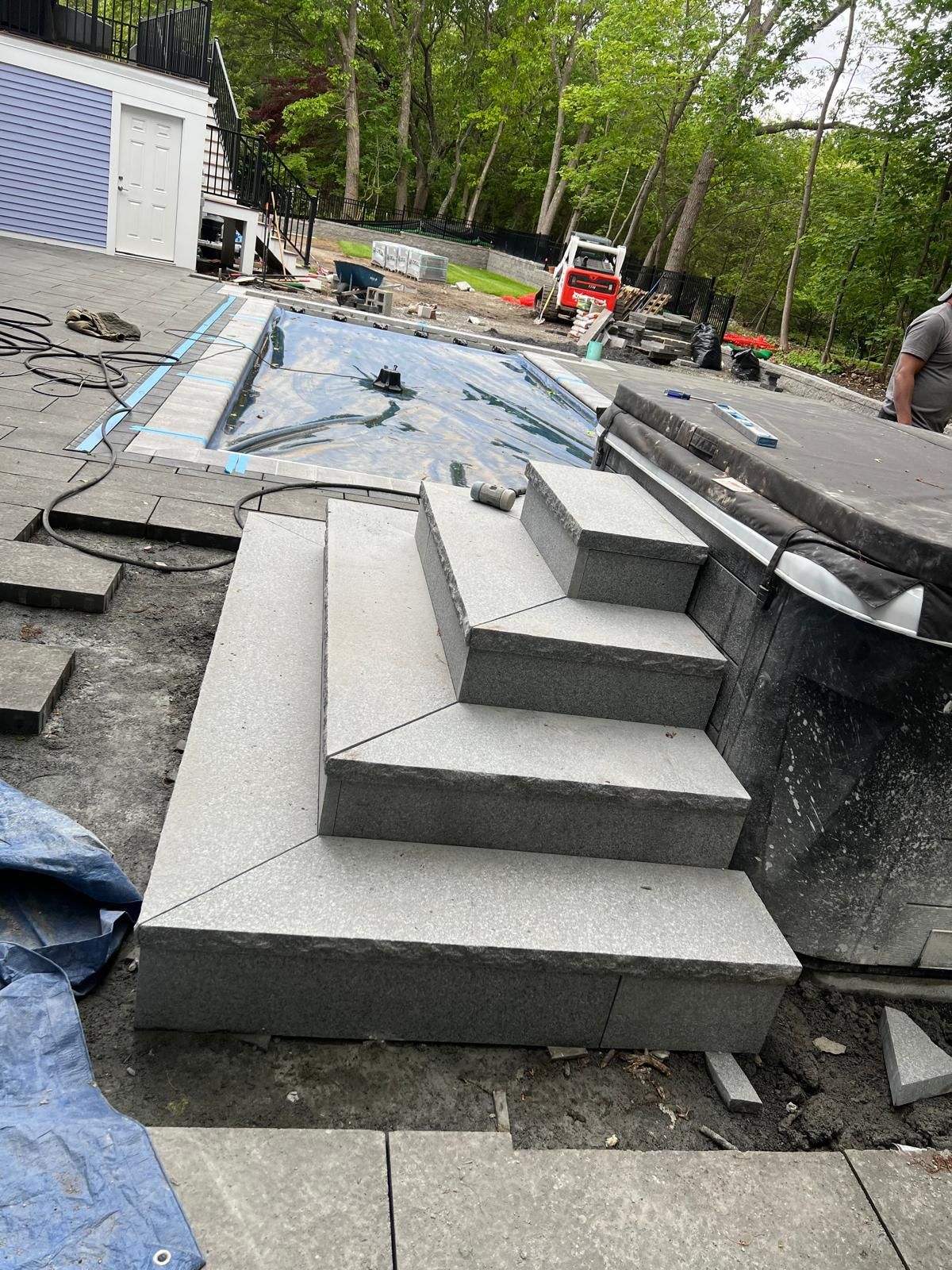 Workers installing a gray stone stairway beside a pool, with construction materials and trees around.