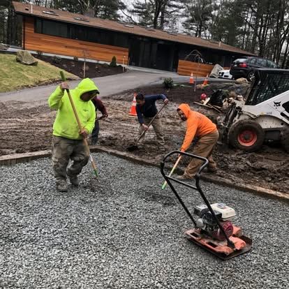 Workers paving a gravel driveway with a plate compactor and tools outside a house in rainy weather