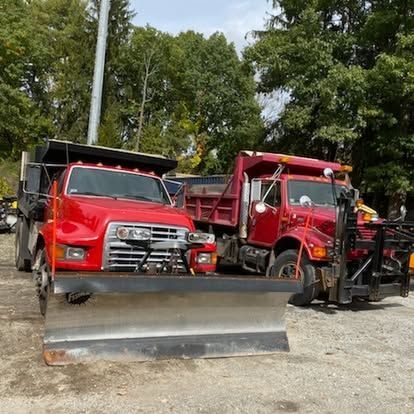 Two red dump trucks parked on gravel behind a metal snowplow blade