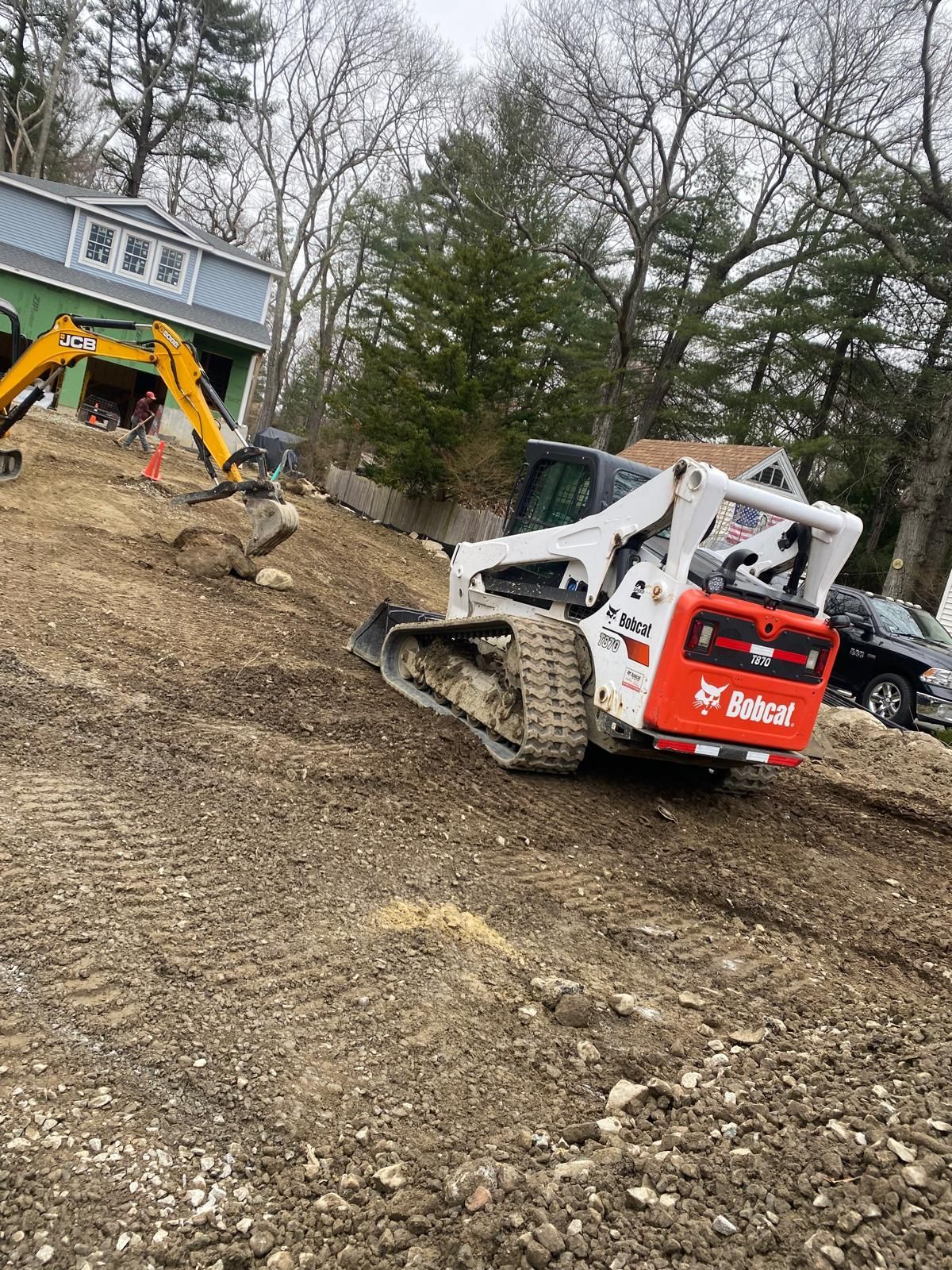 Two skid steer loaders on a muddy hillside near a house, one orange and one white.