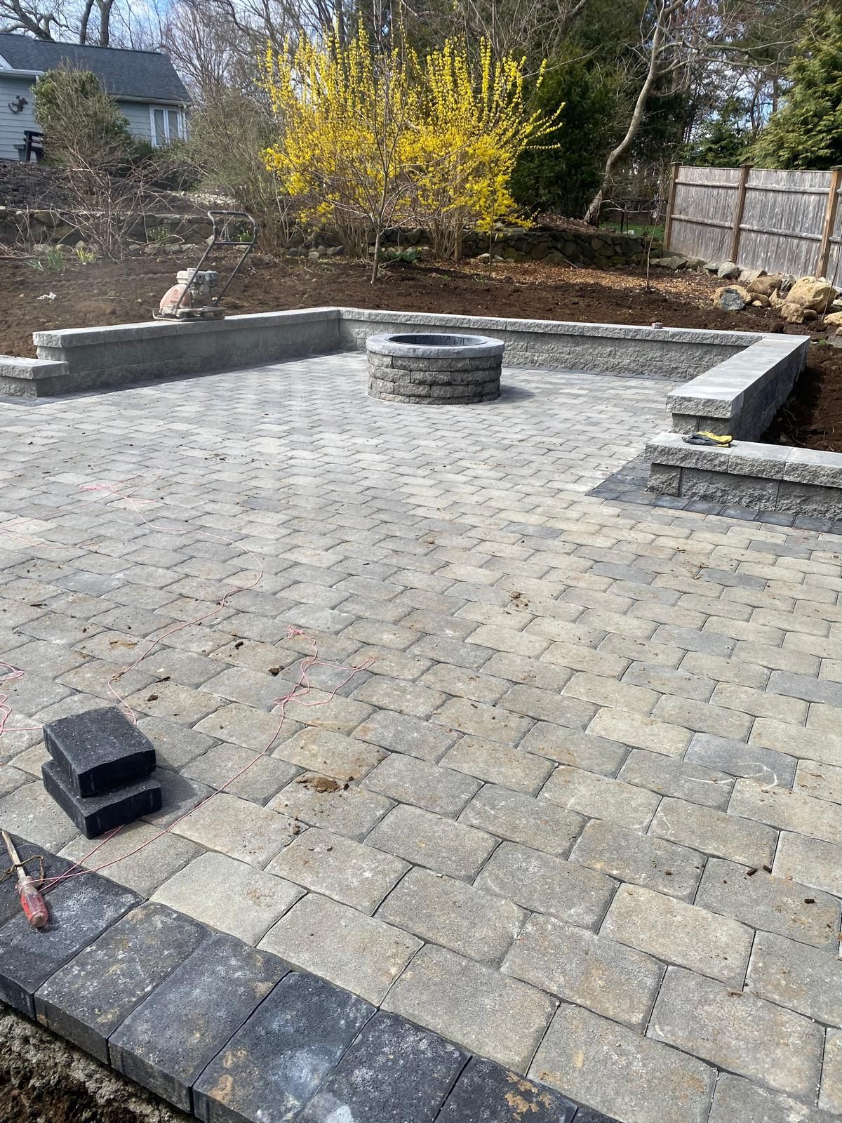 Empty paved patio with low stone walls and steps, bordered by trees and shrubs in a backyard.