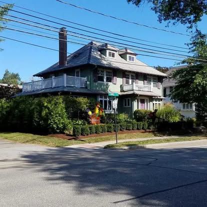 Large gray house with wraparound porch and chimneys on a sunny residential street