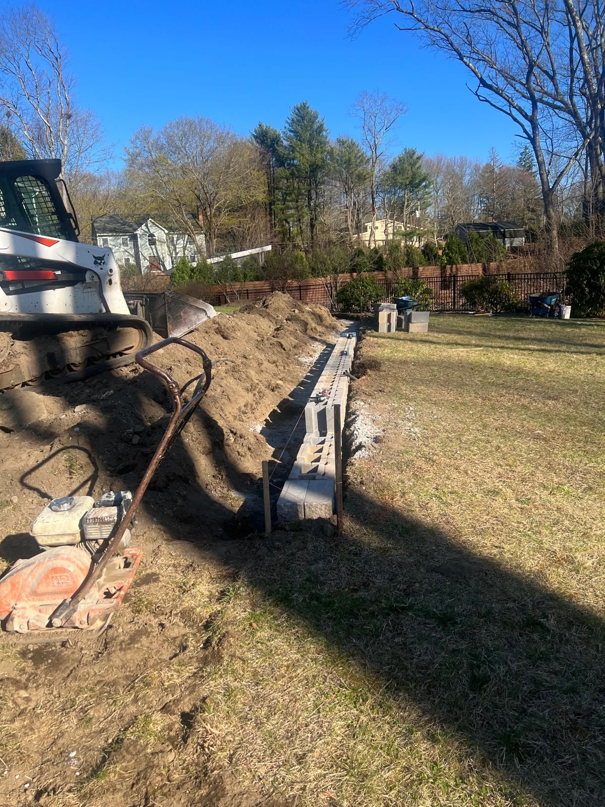 Construction site with excavator, dirt trench, and stacked concrete curbing beside a grassy yard