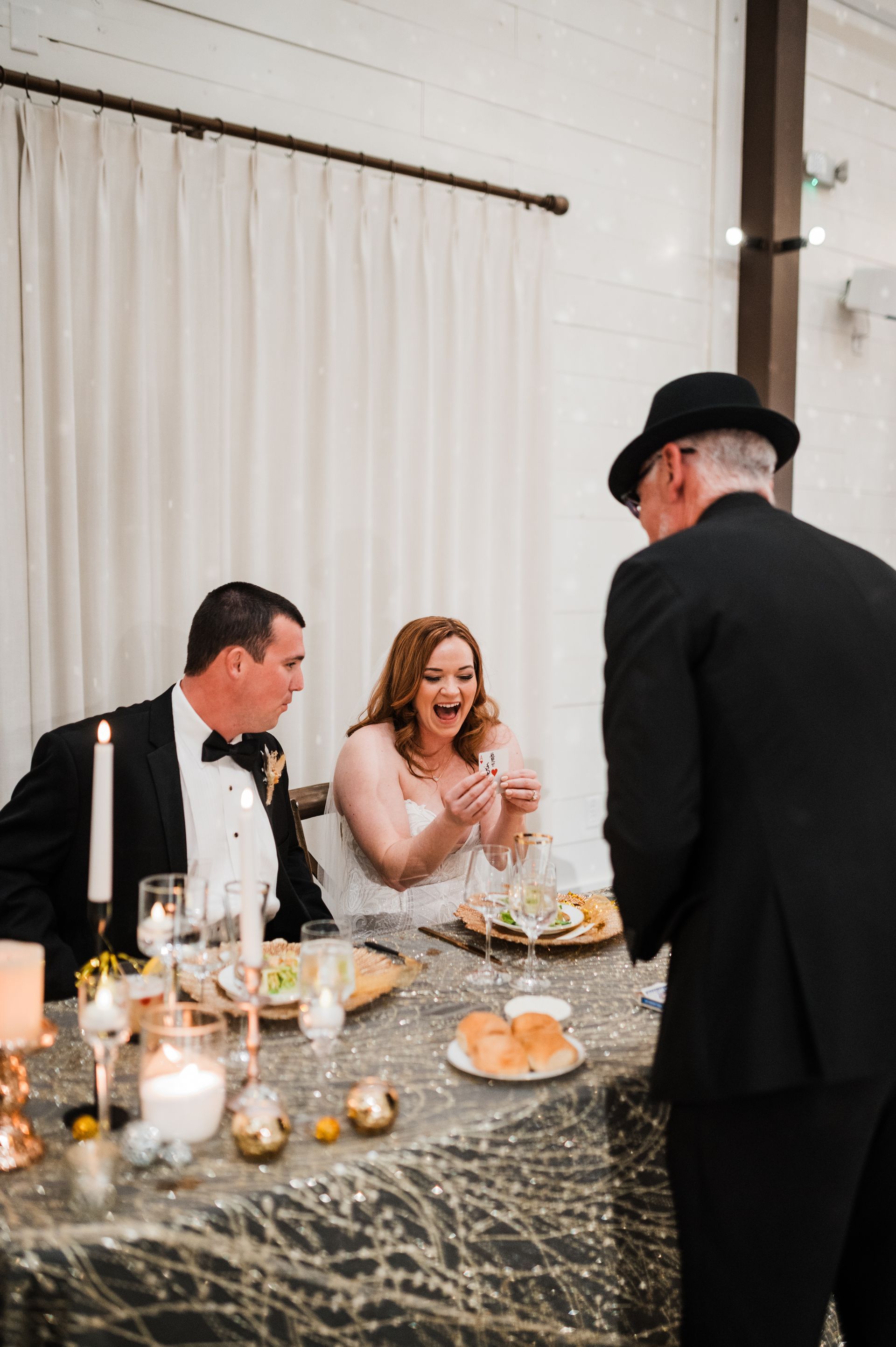 A bride and groom are sitting at a table with a man in a hat.
