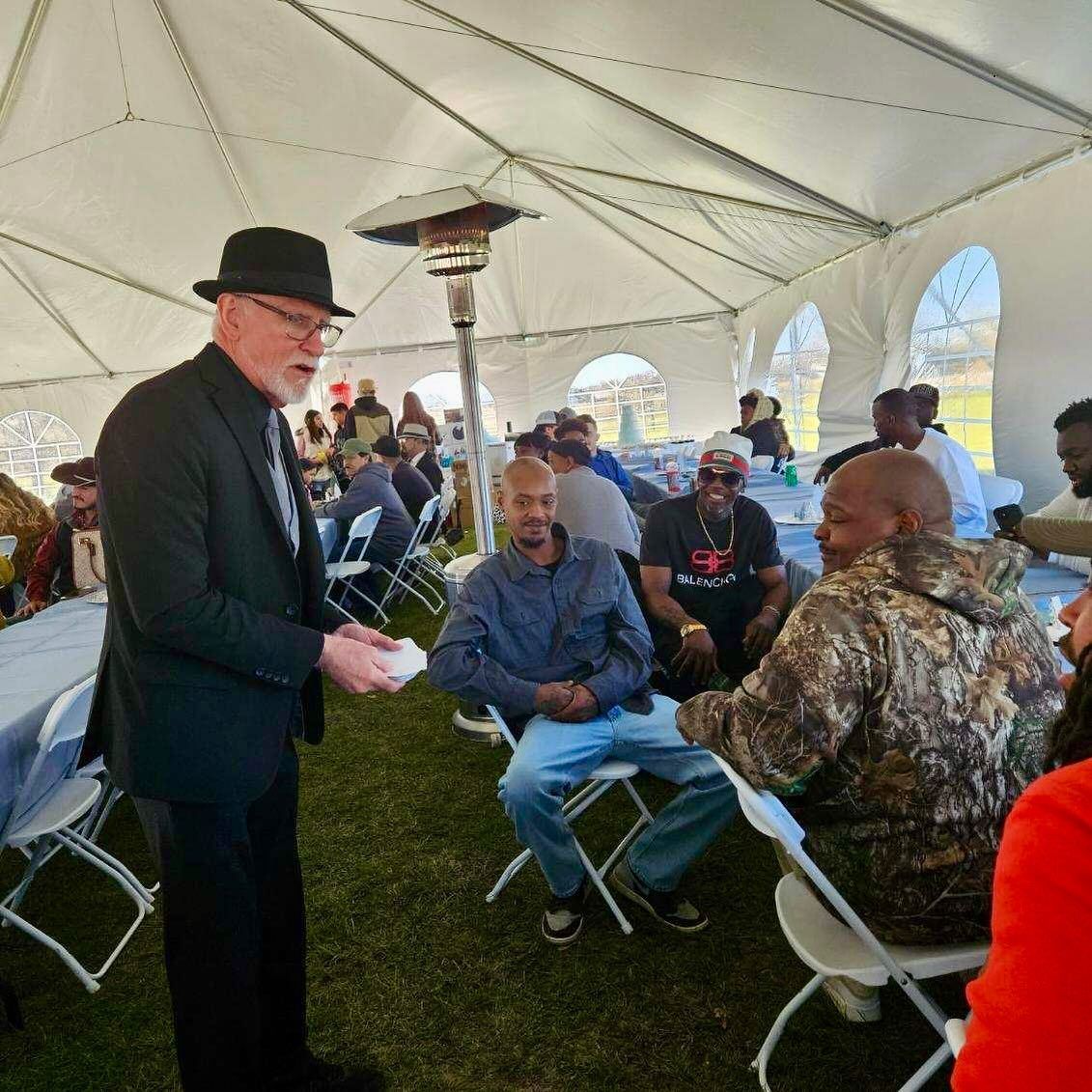 A man in a suit and hat is standing in front of a group of people under a tent.