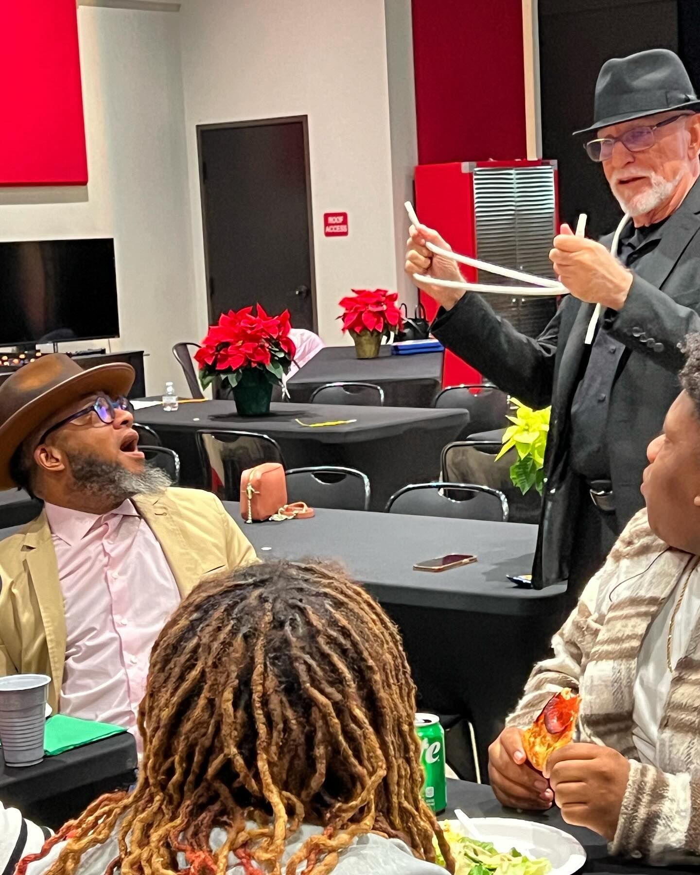 A man in a hat is holding chopsticks in front of a group of people sitting at tables.