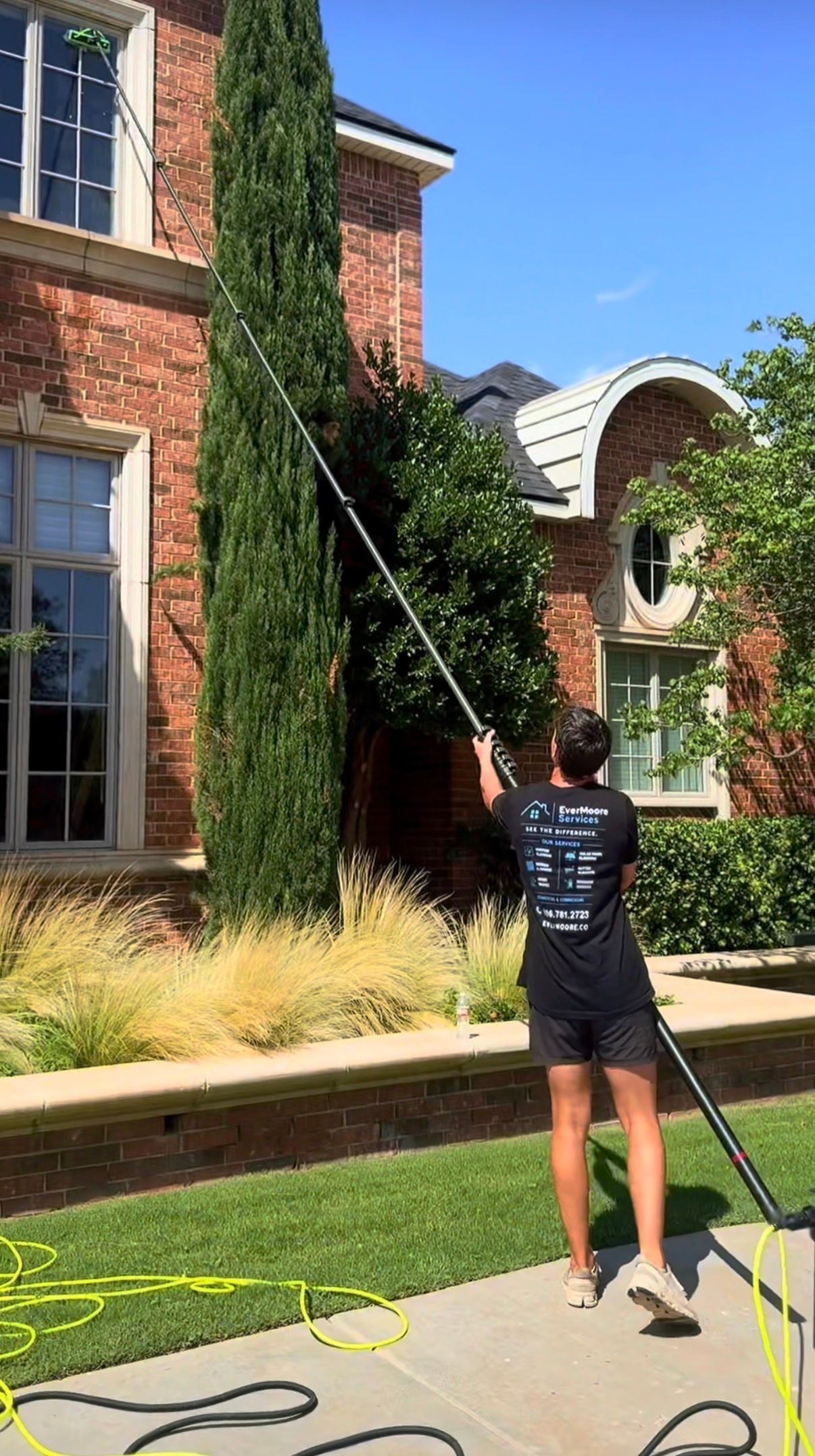 Person using a long pole to reach ivy on a brick building in a sunny courtyard.