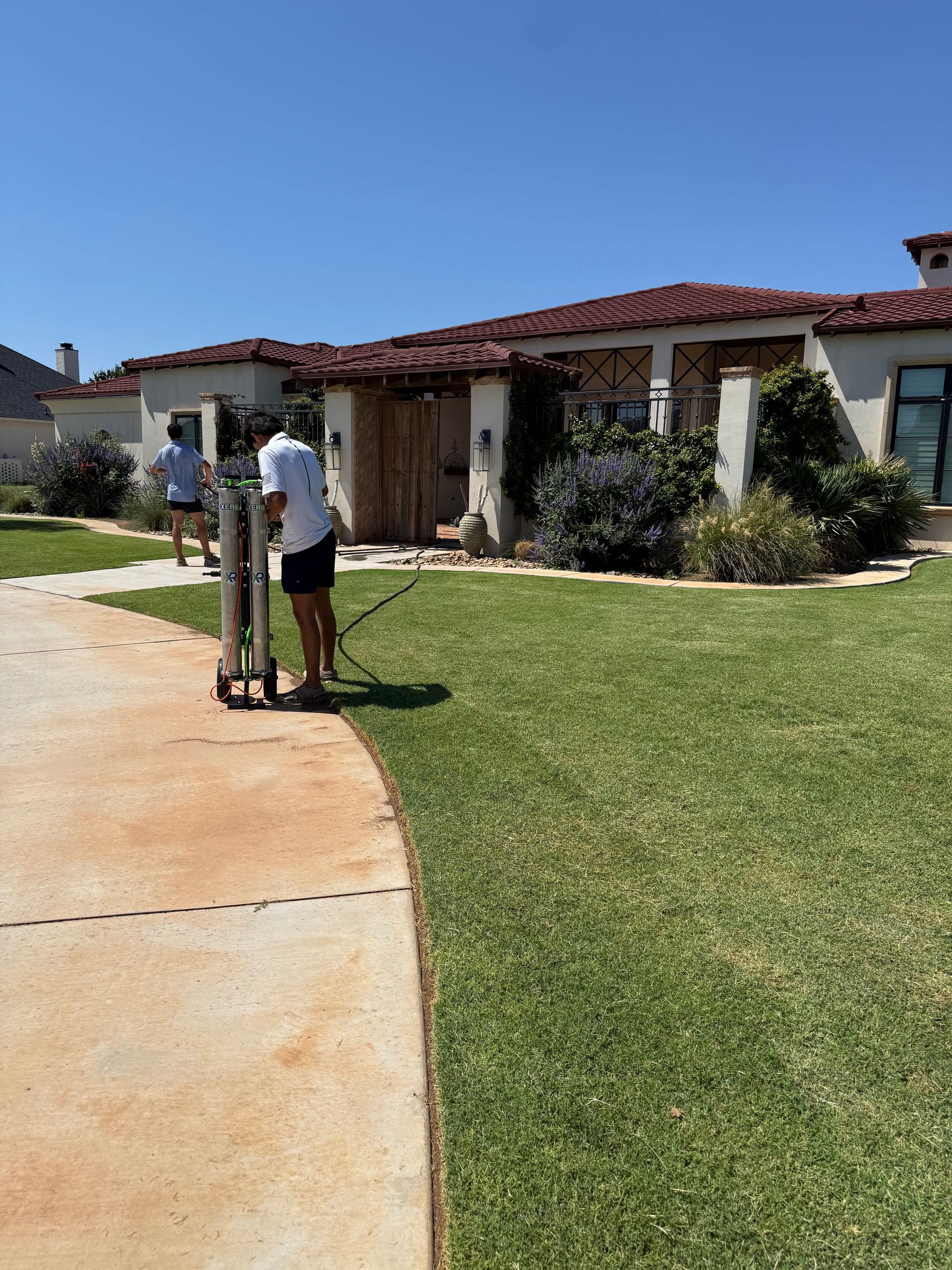 Two people standing on a sidewalk near a stucco house with a red tile roof on a sunny day