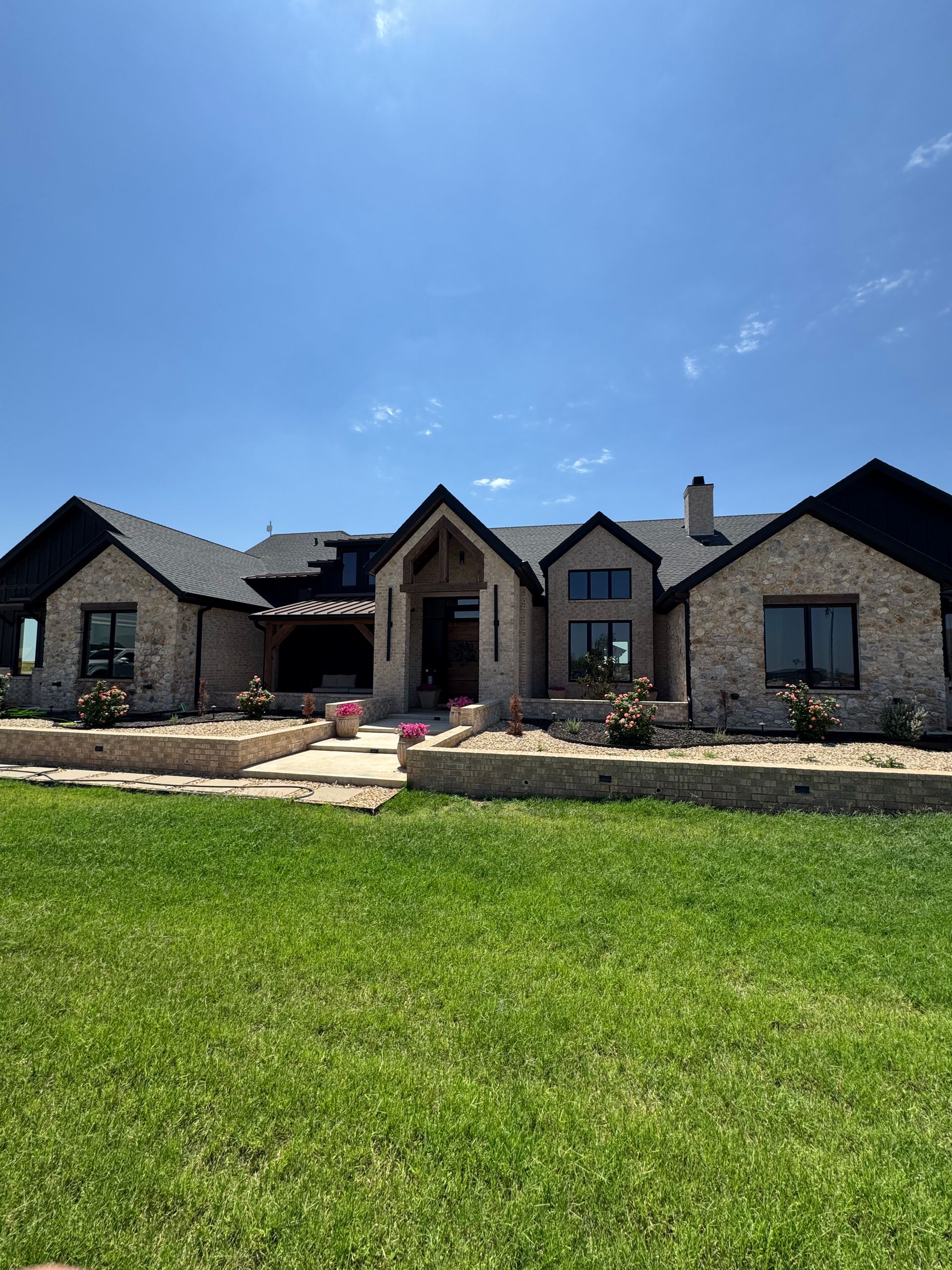 Stone house with a dark roof and front porch, set behind a green lawn under a bright blue sky.