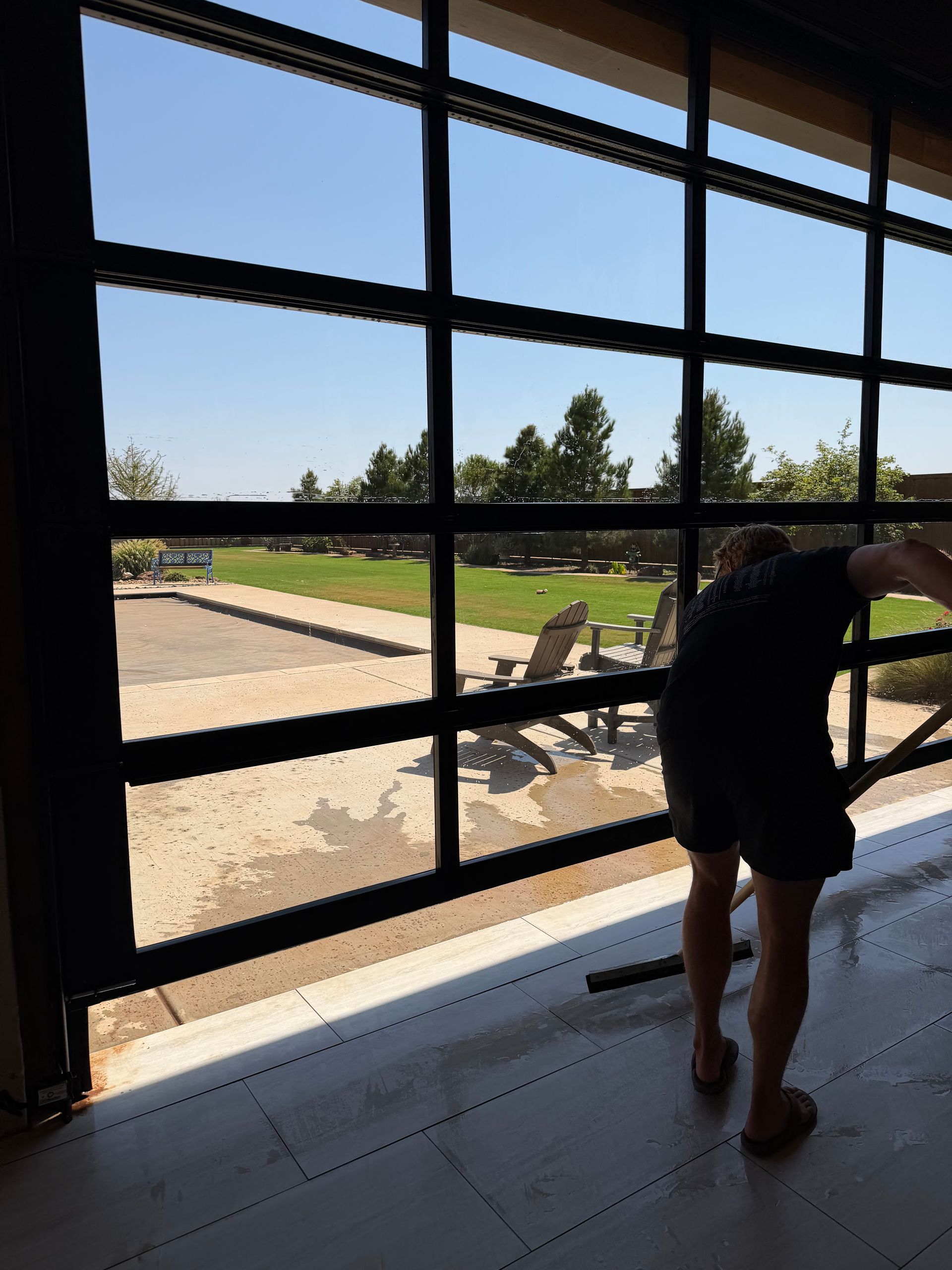 Person looking out a large window at a sunny outdoor courtyard with tables and chairs.