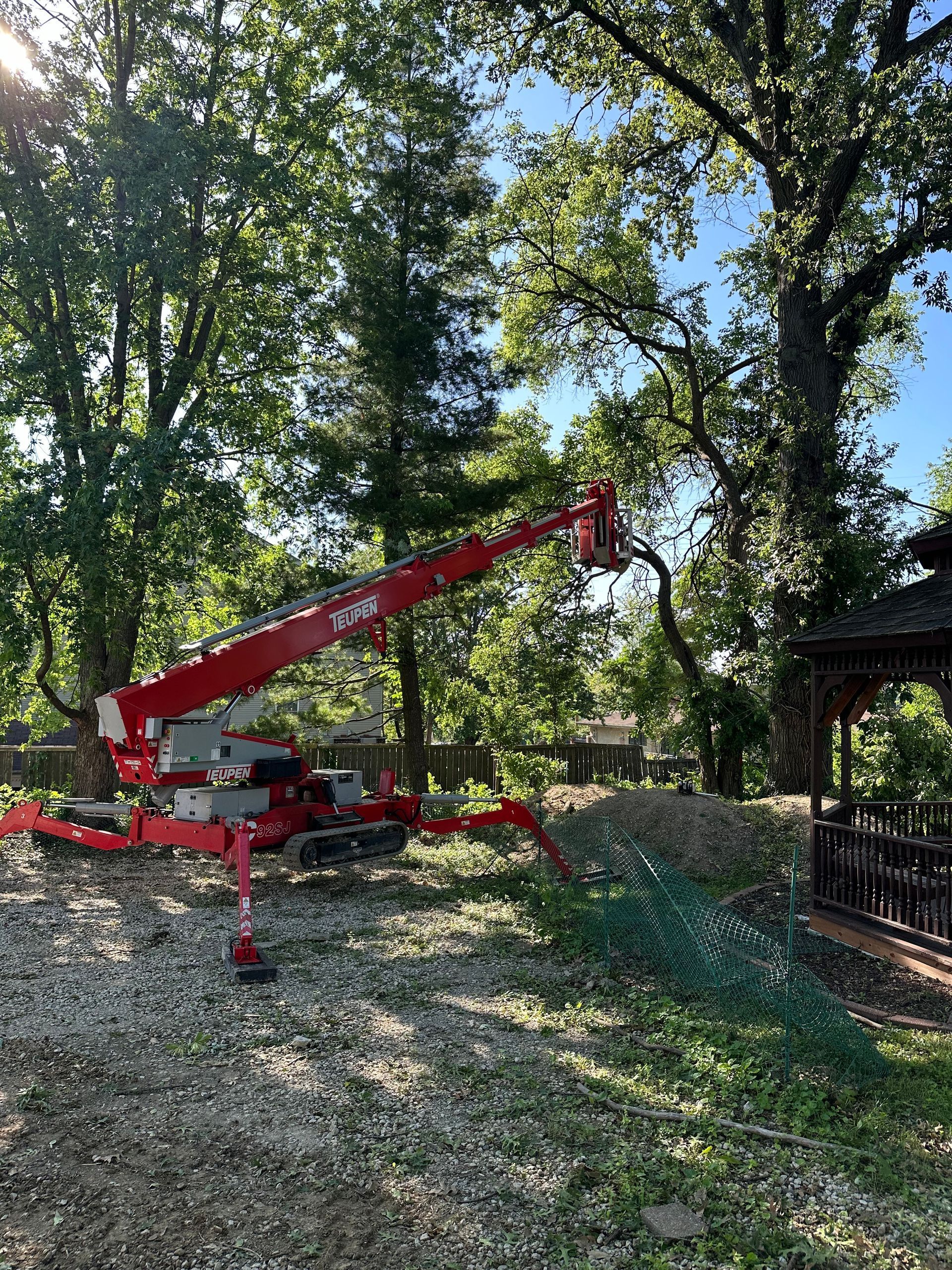 Red tree lift trimming branches, near a gazebo and trees, on a sunny day.