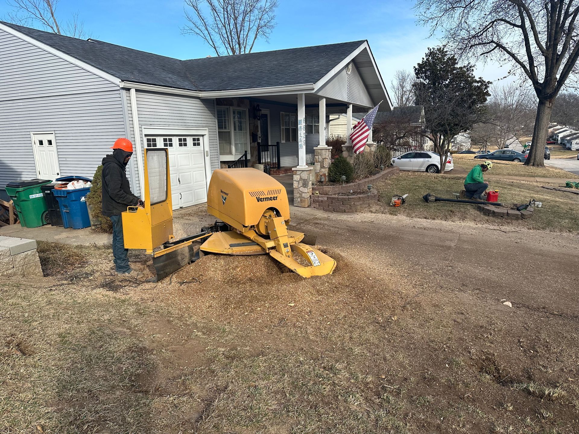 A tree stump is being ground down with a yellow stump grinder in front of a house. Two workers are present.
