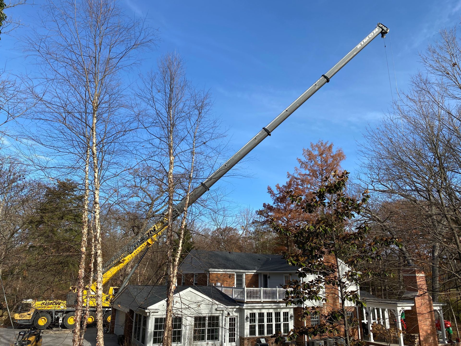 Crane arm over a house, removing trees. Blue sky, fall foliage, and yellow machinery.