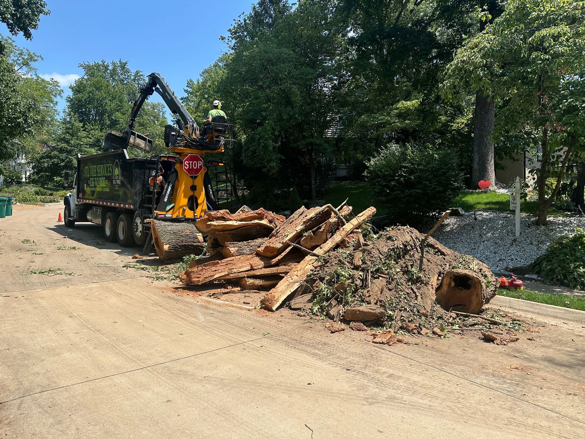 Tree removal in progress: wood pieces piled on the road being loaded into a truck by a machine with a worker on top.