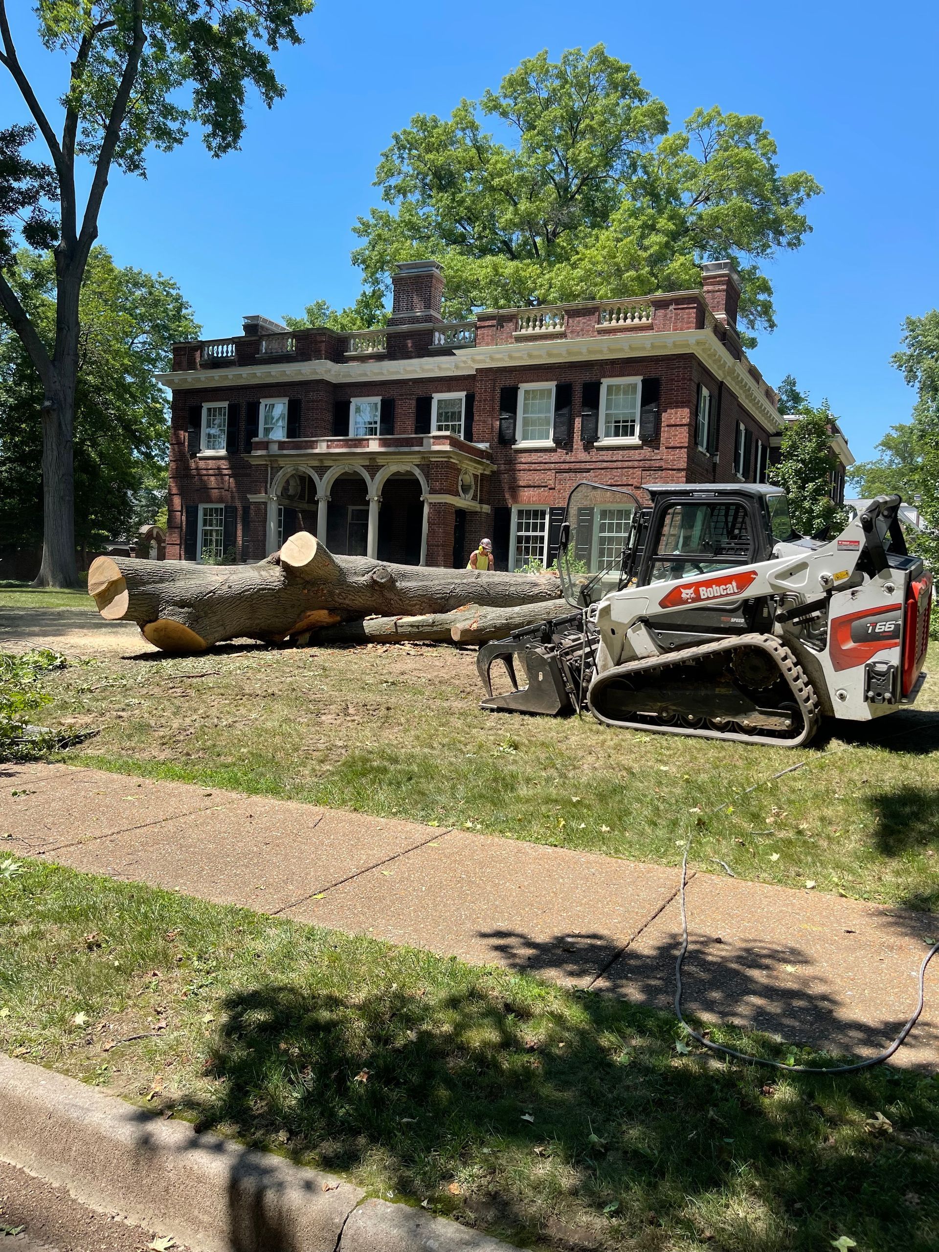 A Bobcat skid-steer loader lifts a fallen tree trunk in front of a large brick house.