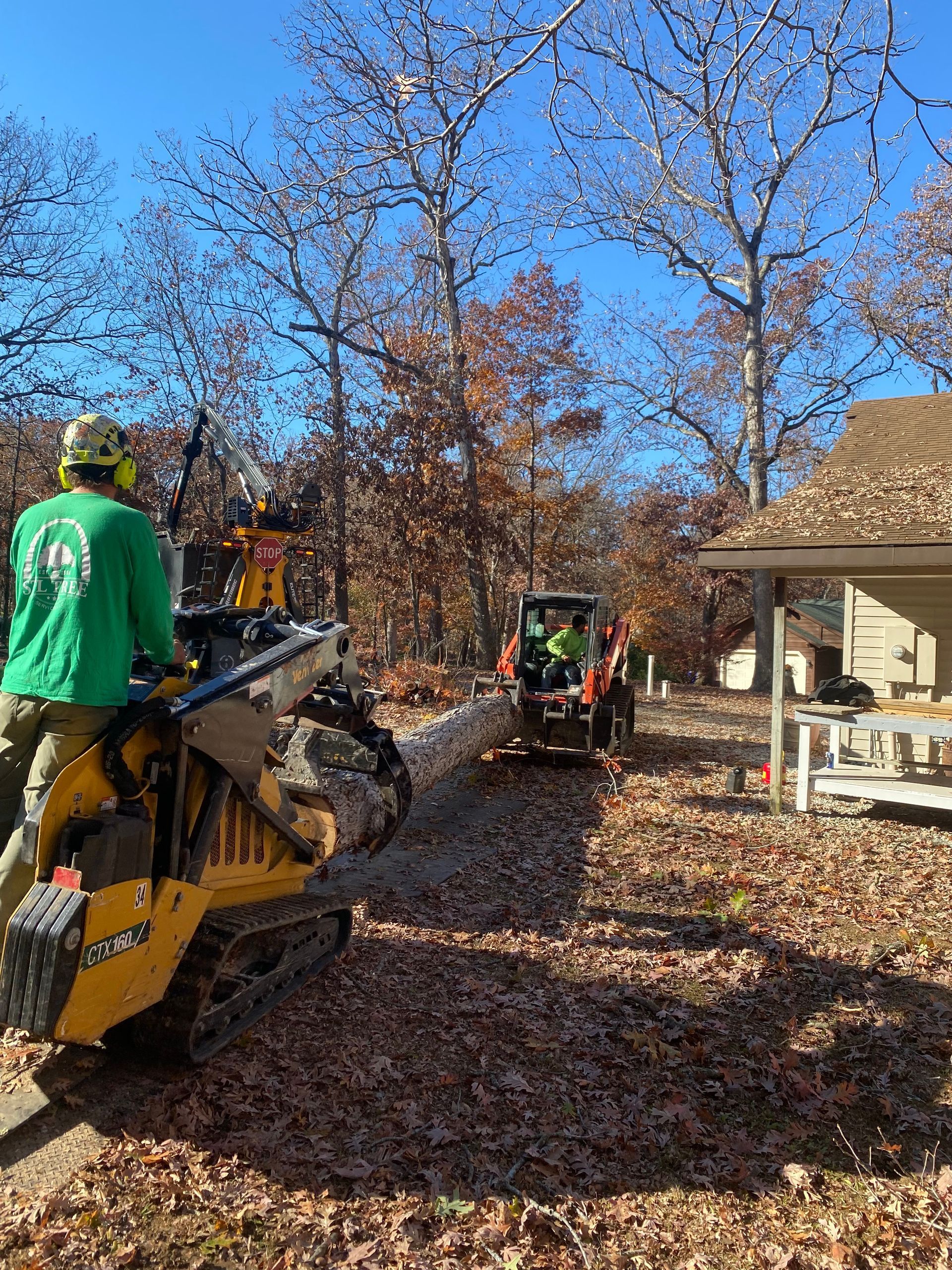 Two workers operate machinery to move a log through a leaf-covered yard; a house is to the right.