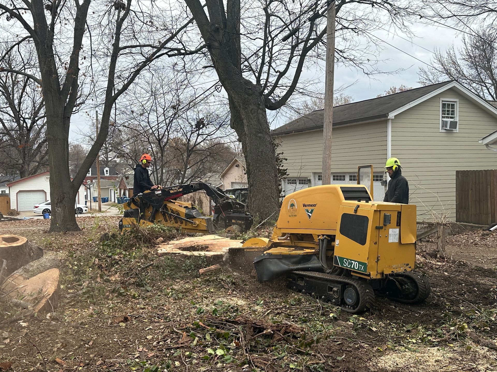 Two people operating stump grinding machines in a yard next to a house.