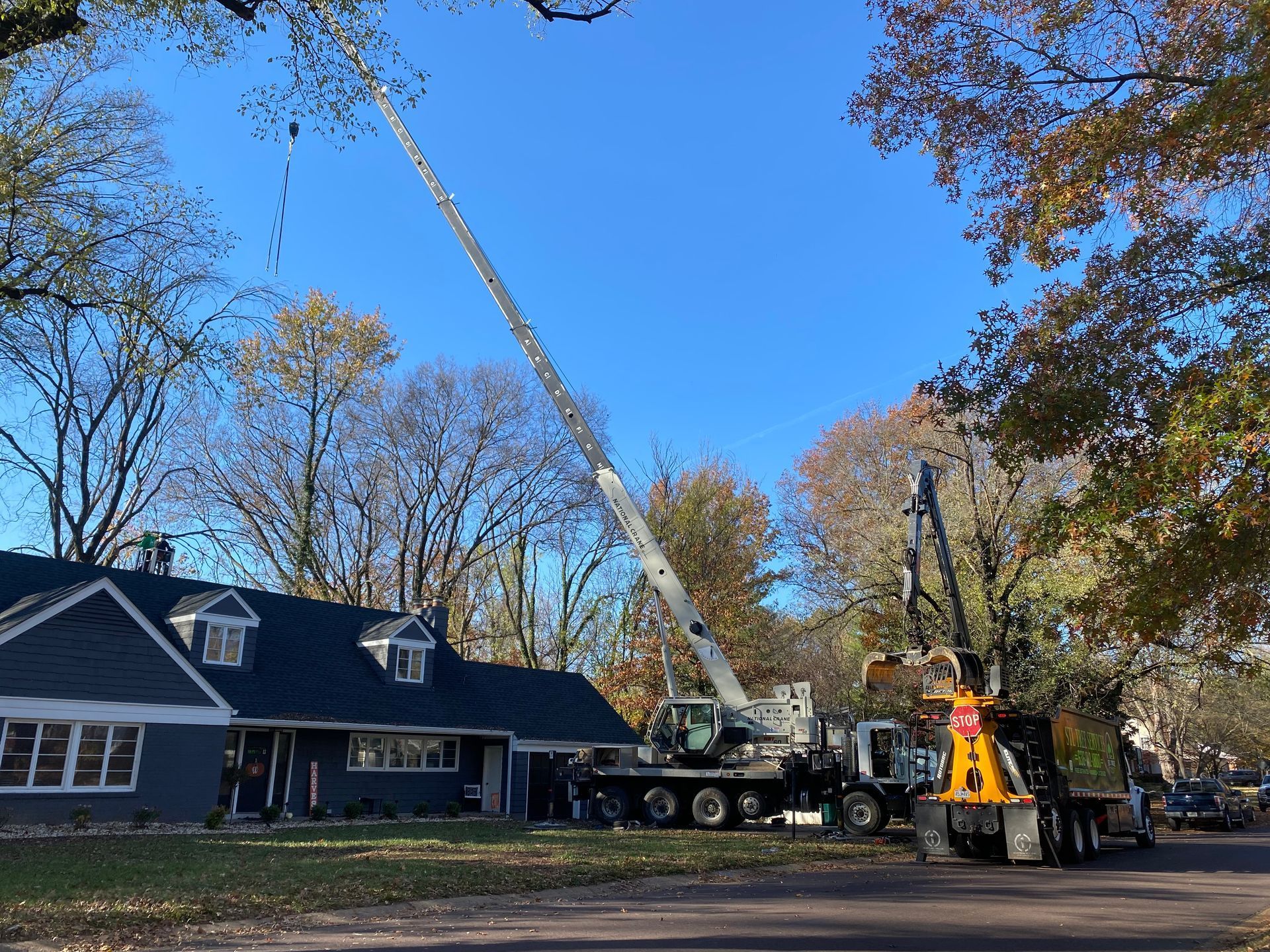 Tree trimming service: Crane cutting branches near a blue house. Chipper truck collects debris; sunny day.