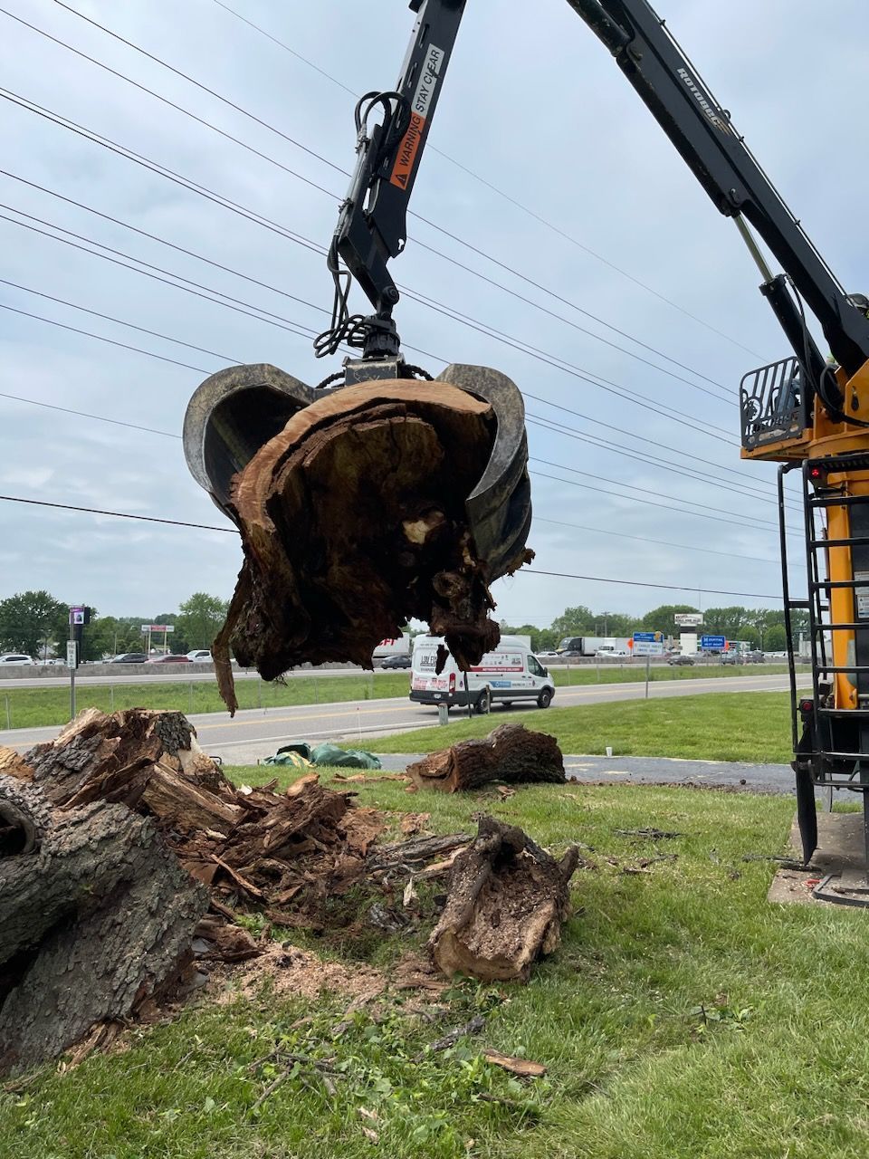 A mechanical arm lifts a large, broken tree trunk. Chunks of wood litter the grassy area near power lines.