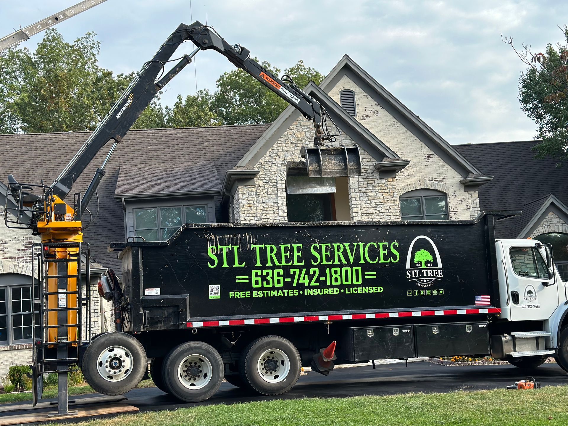 Tree service truck with crane trimming trees near a house; black and green logo on truck.