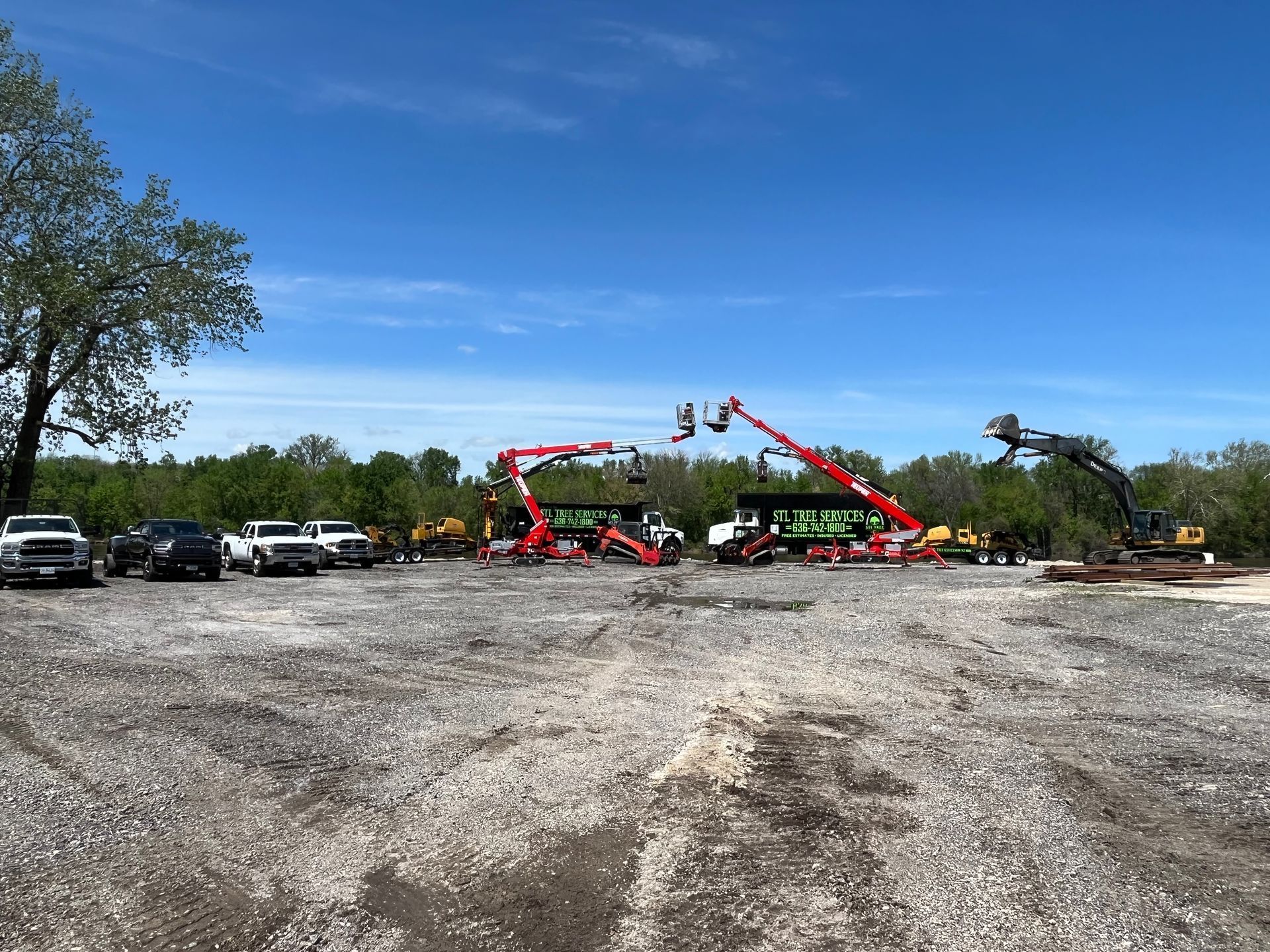 Construction equipment parked on a gravel lot under a blue sky.  Two red boom lifts are raised.