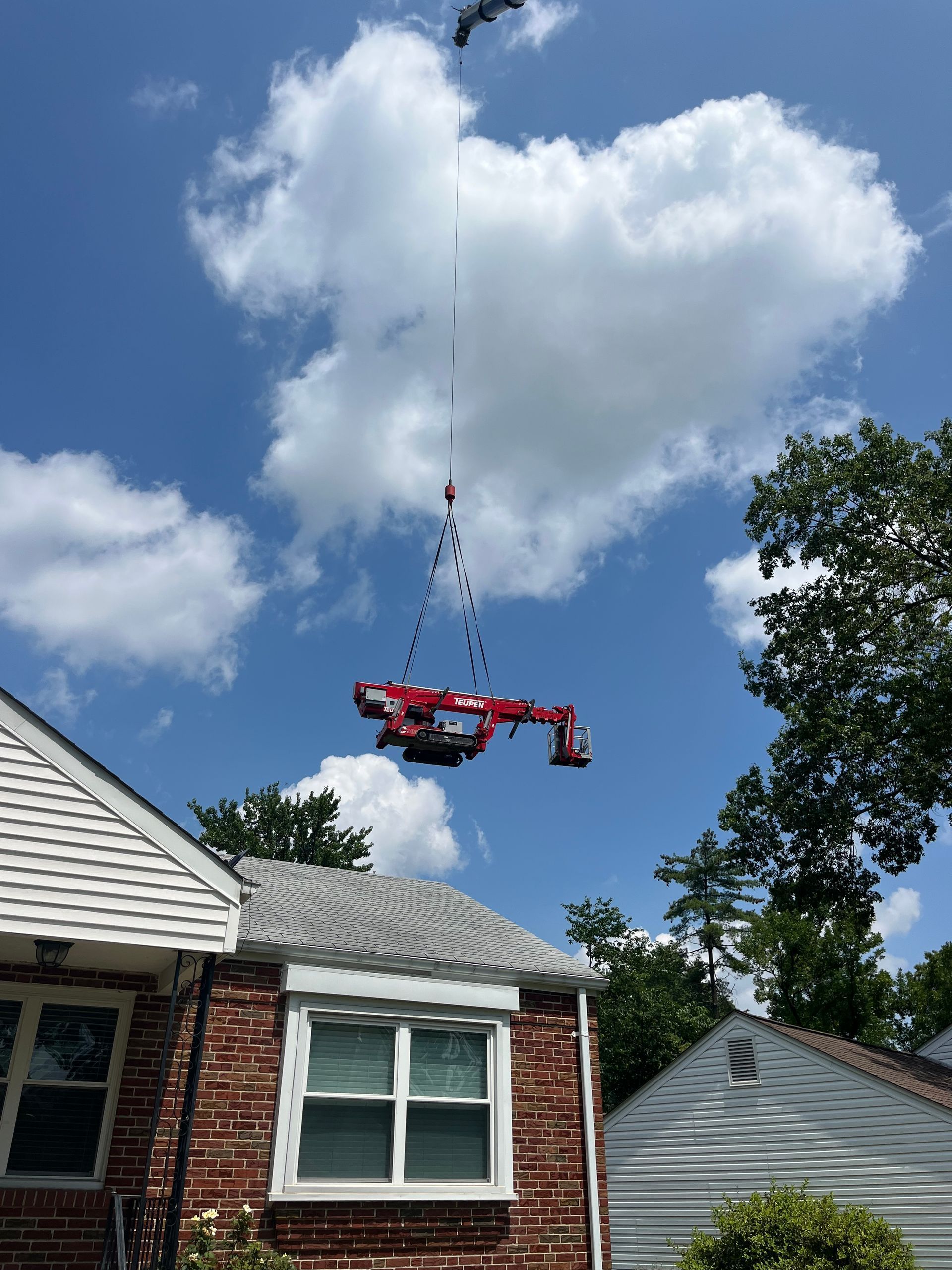Crane lifting a red object over houses against a blue sky with clouds.