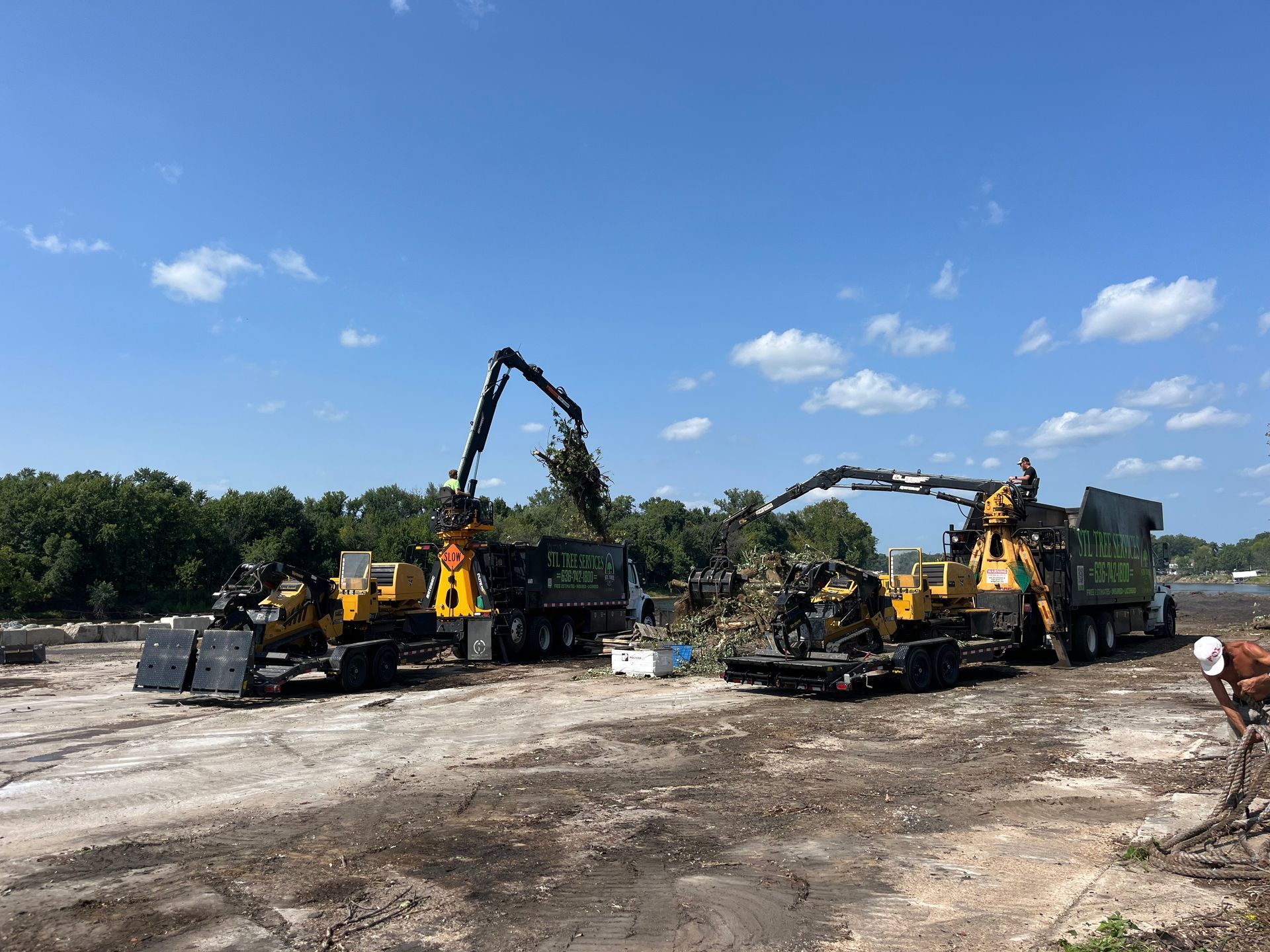 Two industrial wood chippers process trees on a cleared lot under a blue sky.