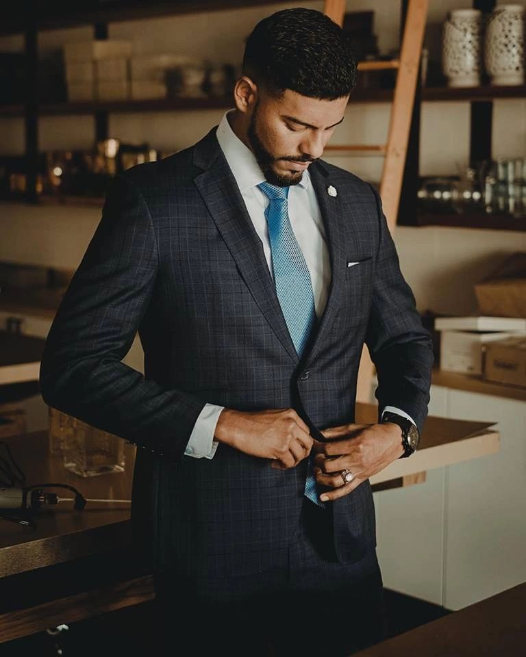 Man in navy suit adjusting jacket button, standing indoors.