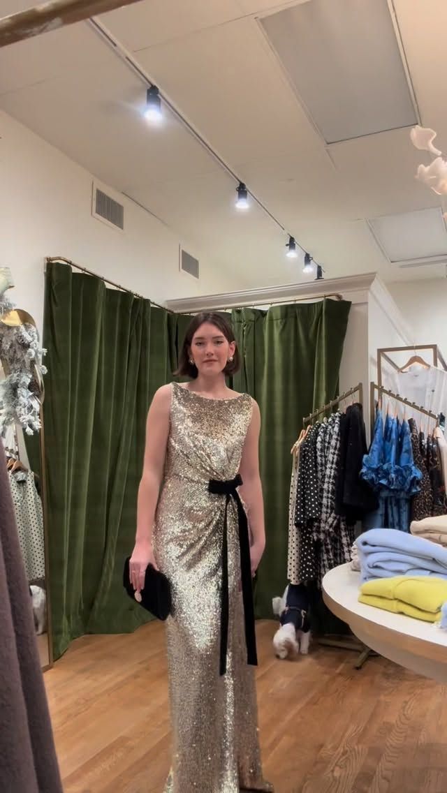 Woman in gold sequined gown, black clutch, bow, standing in a boutique with green velvet curtains.