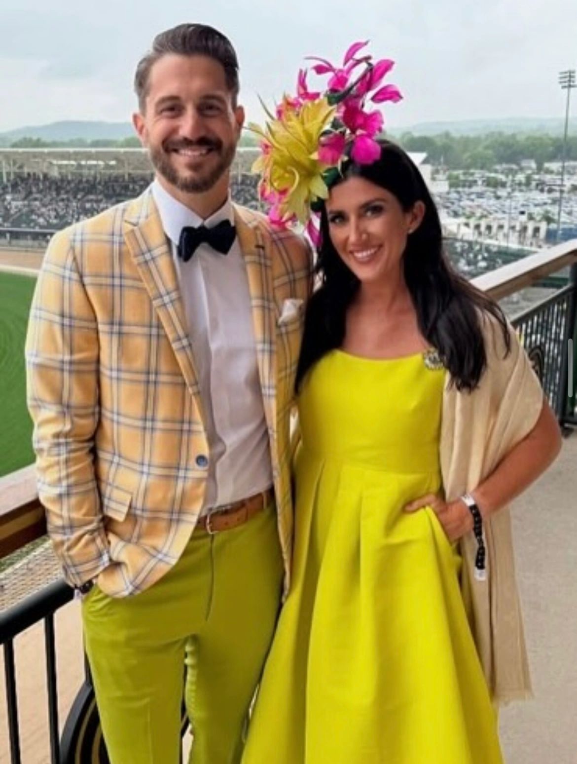 Couple posing at a race track: man in yellow plaid blazer, woman in yellow dress with large floral hat.