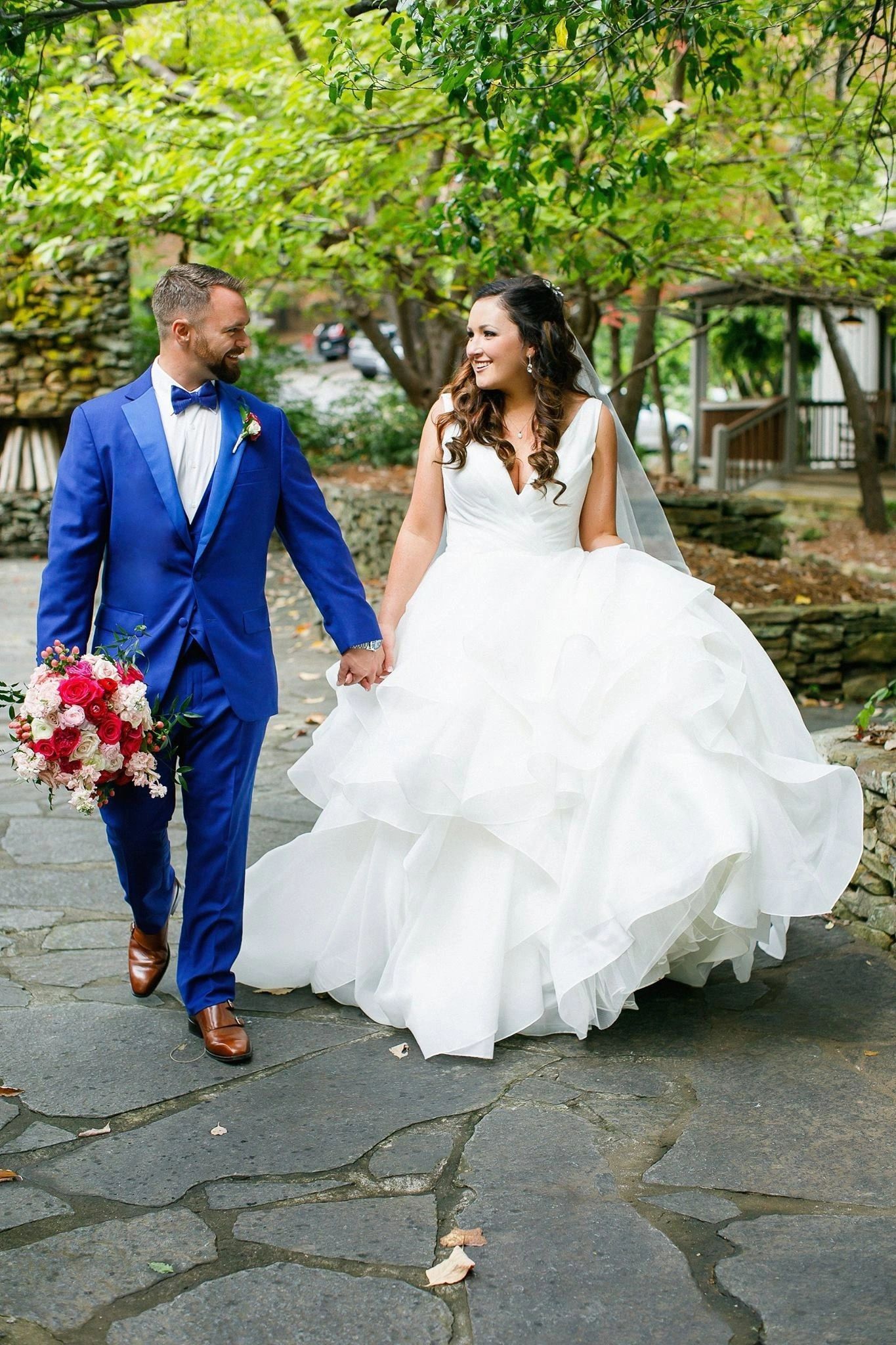 Bride and groom holding hands, walking outside. The groom wears a blue suit, holding a bouquet, and the bride wears a white gown.