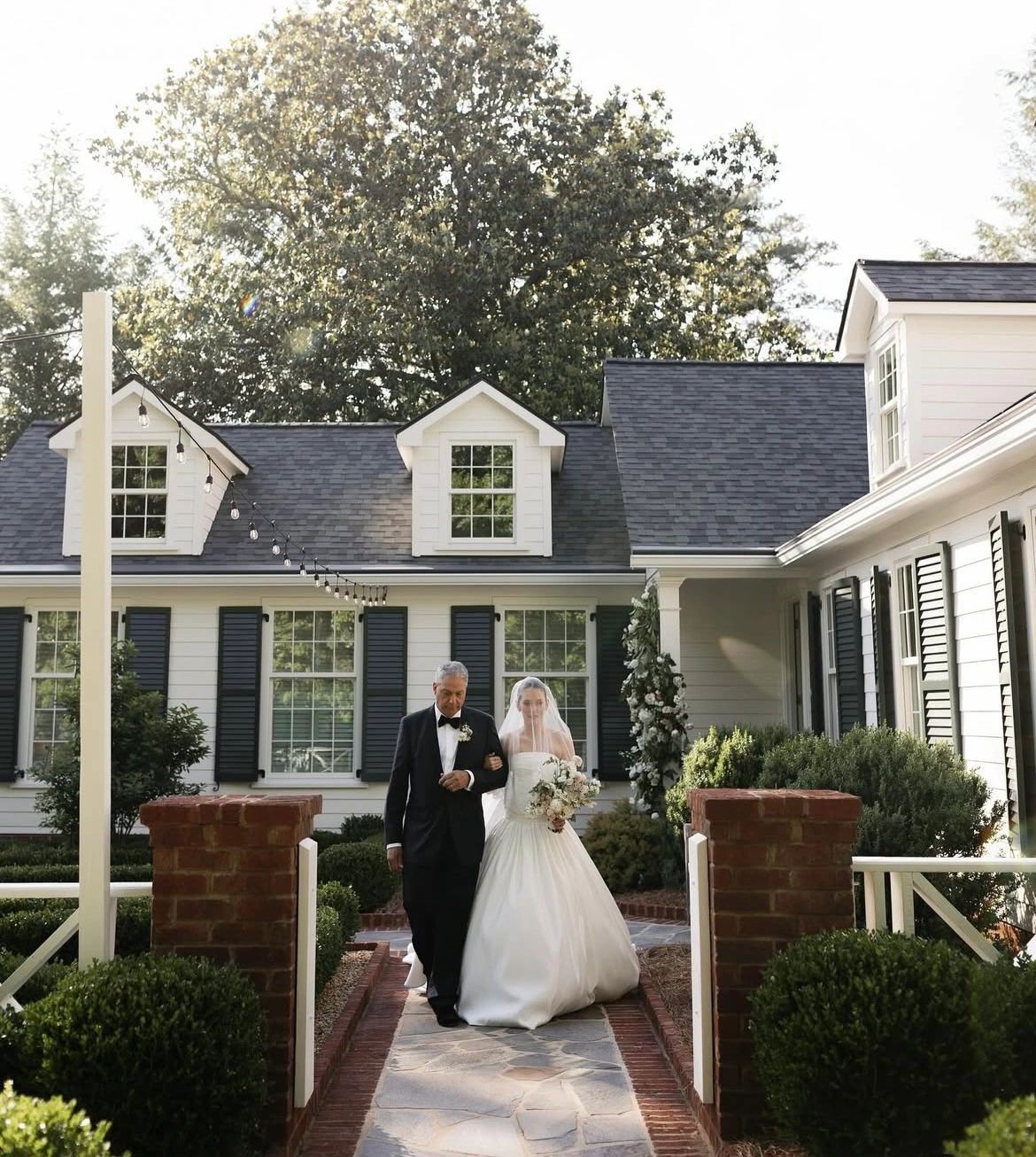 Bride and her father walk toward a white house. The bride wears a white gown. The setting is outdoors.