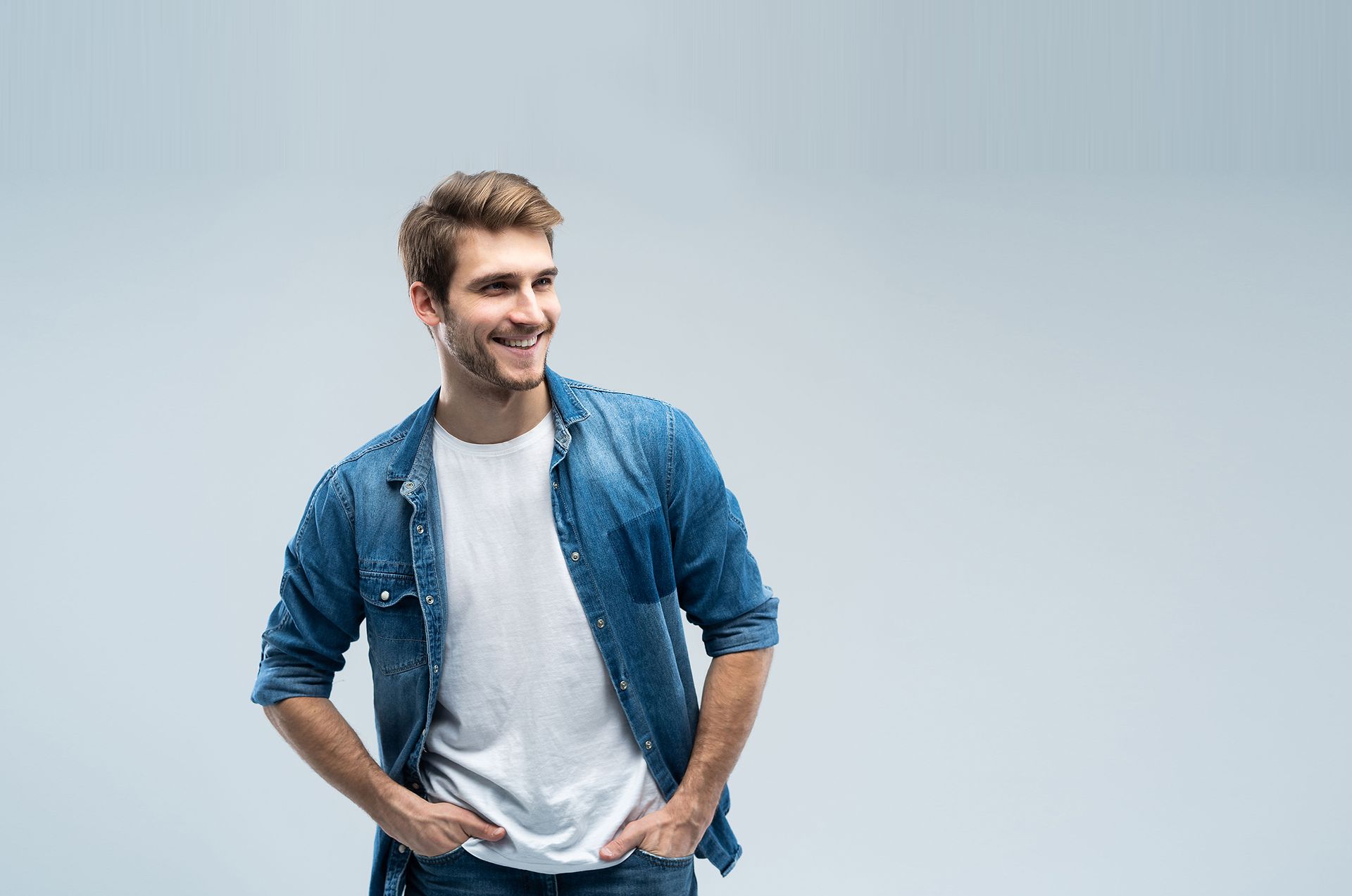 Man smiling, wearing a denim shirt over a white tee, hands in pockets.