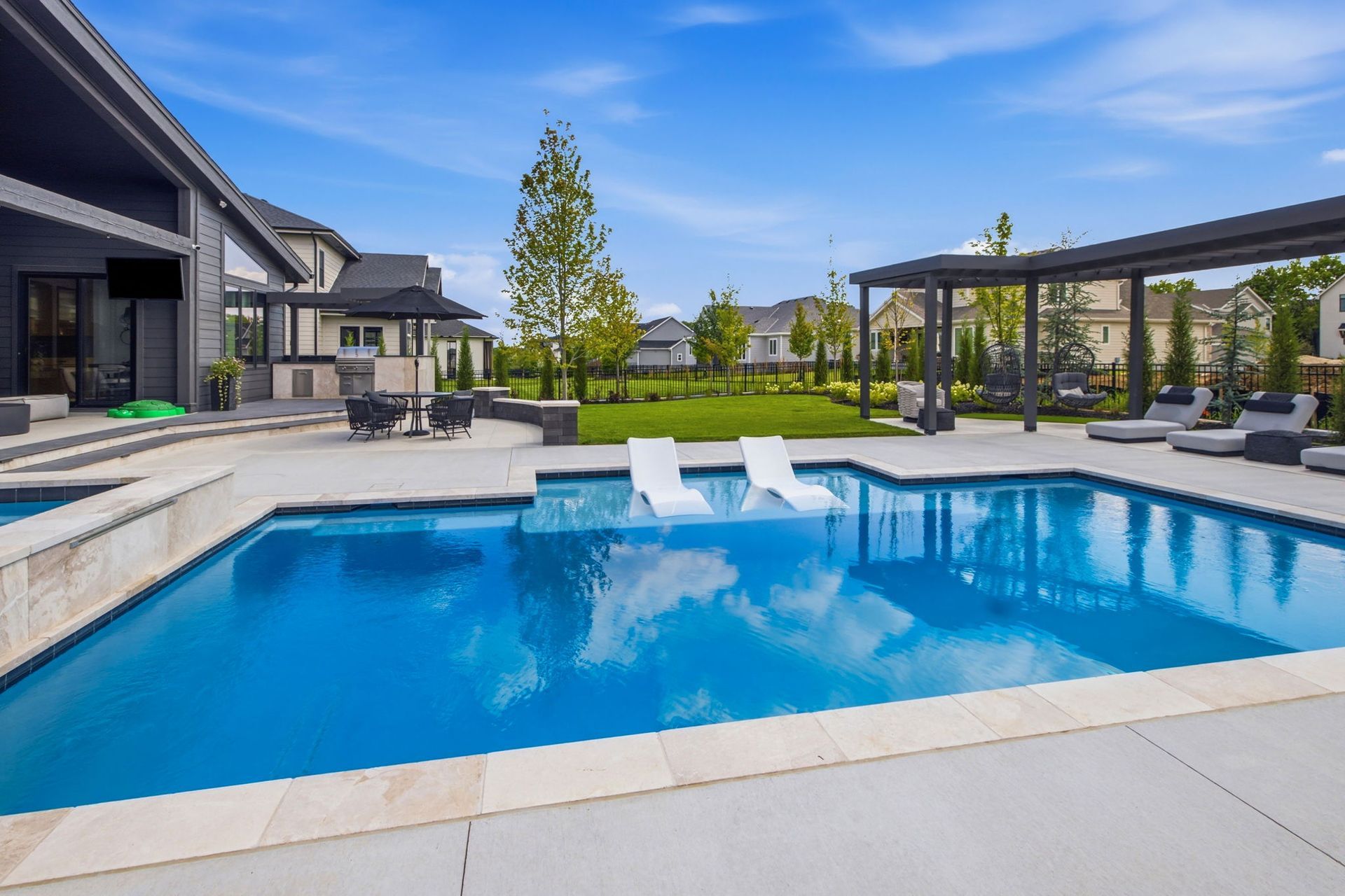 Modern backyard with a blue swimming pool, white loungers, and a covered patio beside a contemporary house