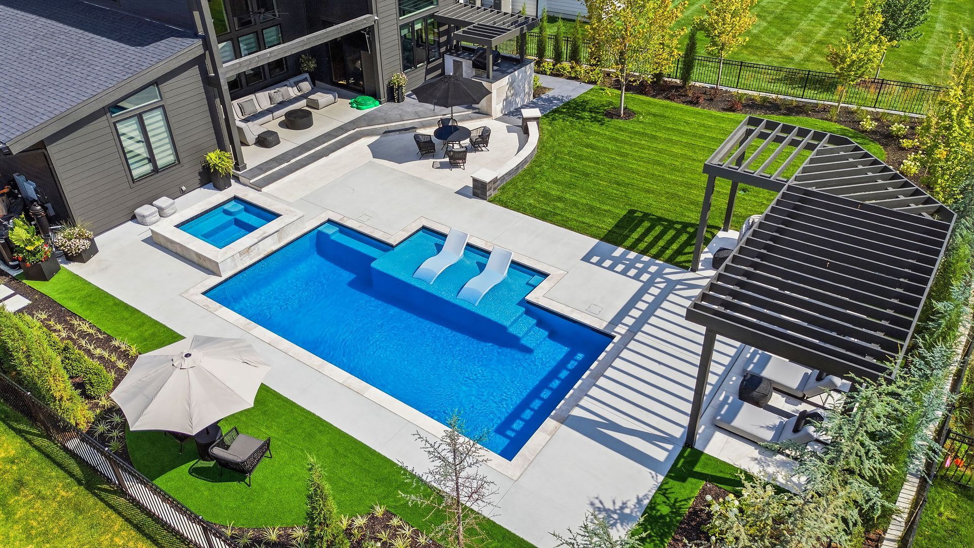 Modern backyard with a blue swimming pool, lounge chairs, patio seating, and a pergola on a manicured lawn.
