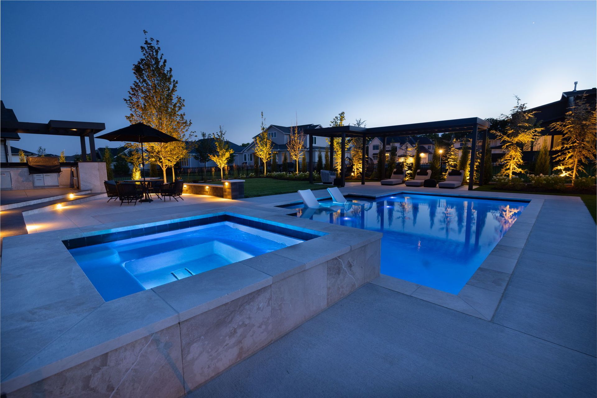 Blue-lit outdoor pool and hot tub at dusk in a modern resort courtyard with loungers and palm trees