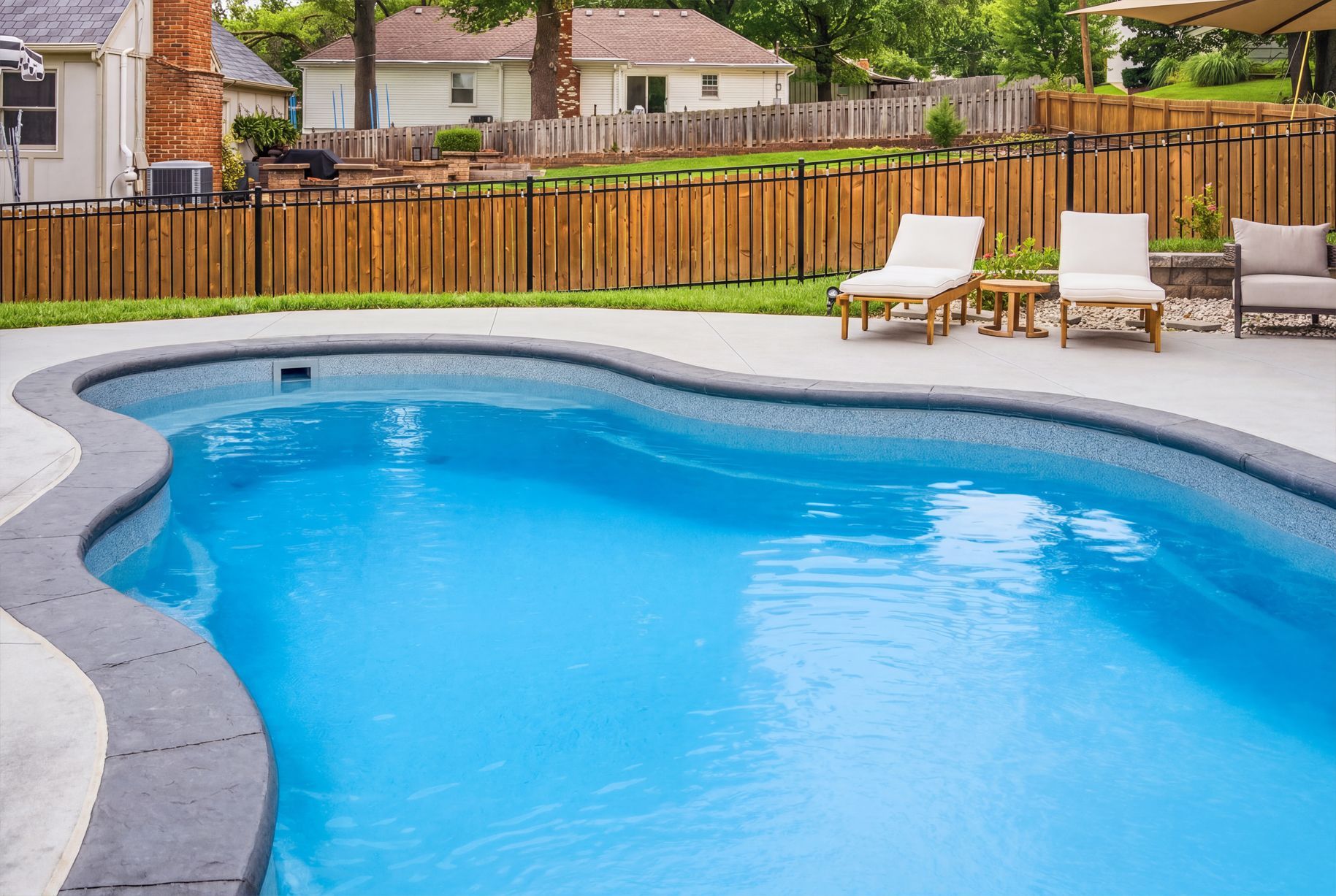 Curved backyard swimming pool with blue water, lounge chairs, and a wooden fence