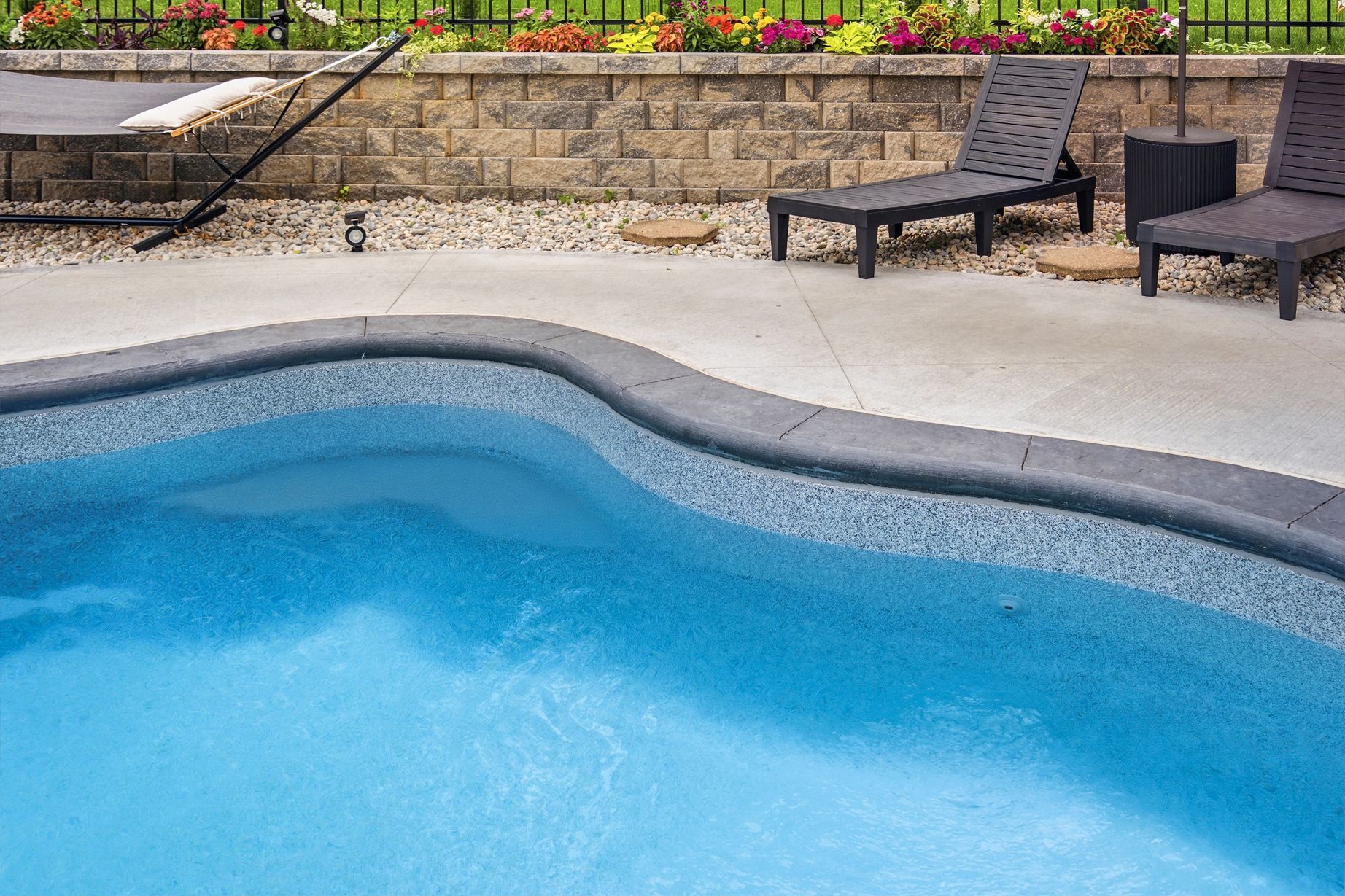 Curved blue swimming pool beside lounge chairs and a stone patio with flowers in the background