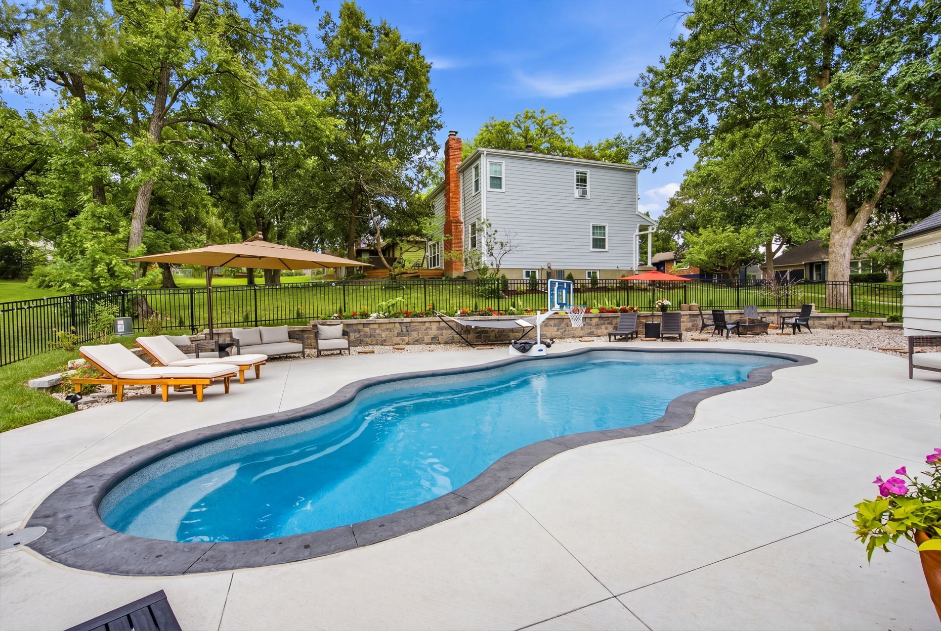 Backyard swimming pool with lounge chairs, patio, and trees under a blue sky