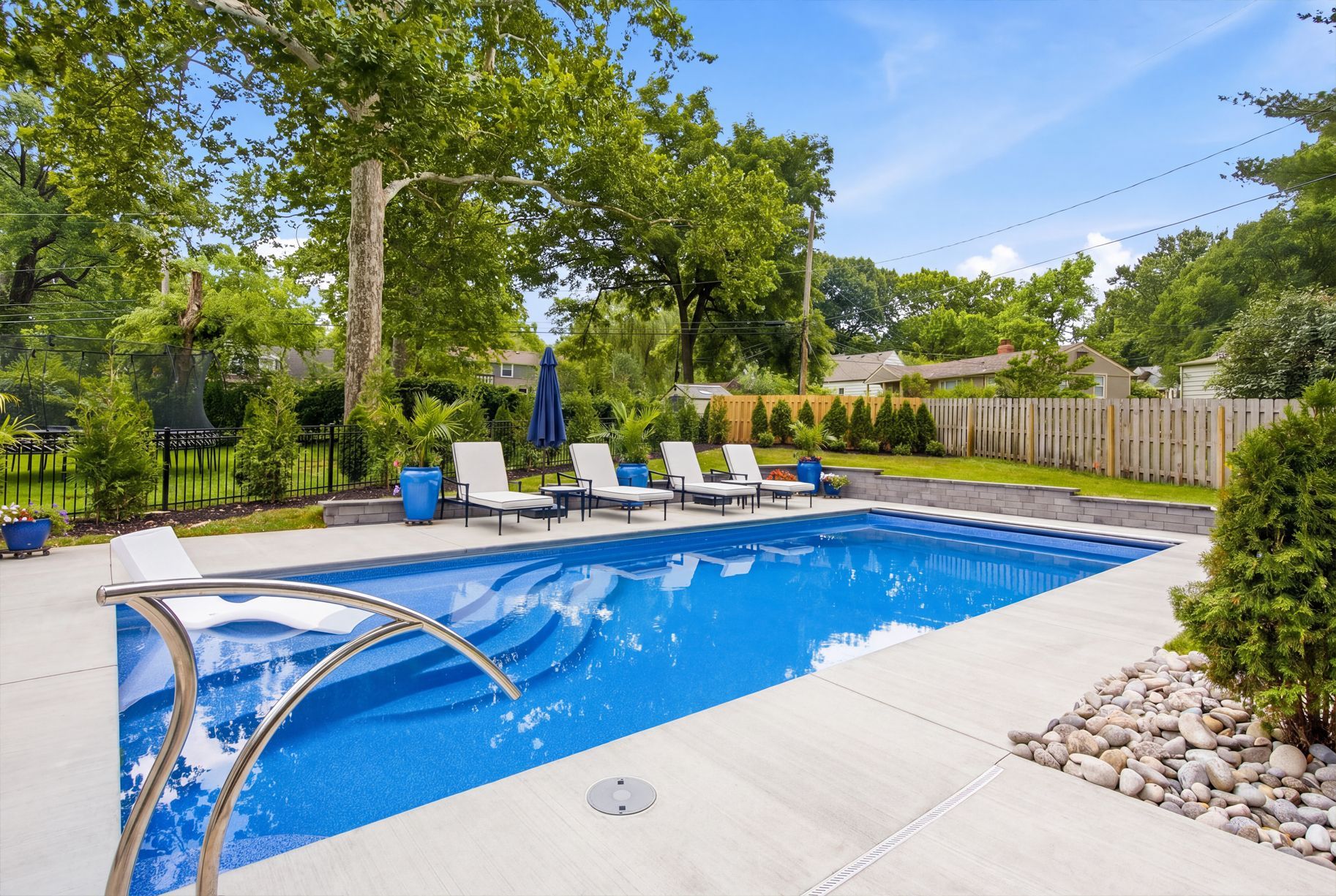 Backyard swimming pool with blue water, lounge chairs, and patio under trees and a fence
