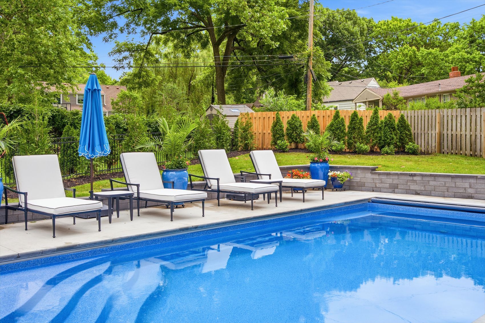 Backyard pool with white lounge chairs, blue umbrella, and green trees behind a fence