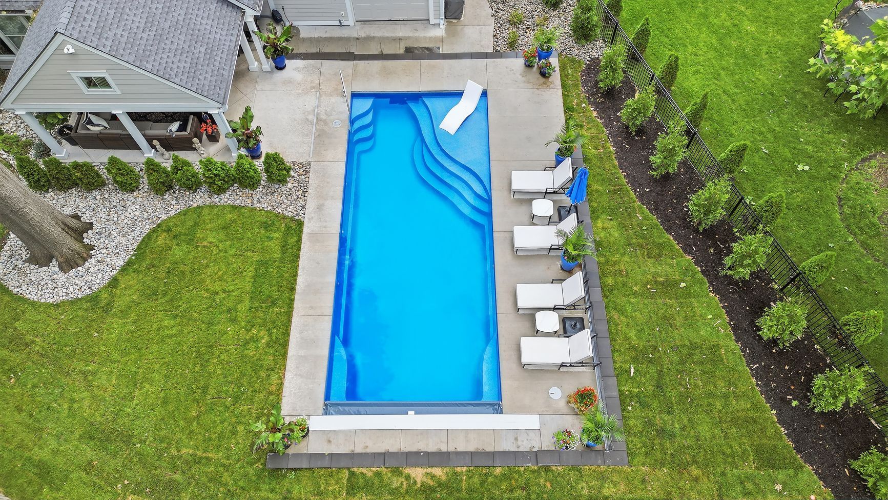Aerial view of a backyard pool beside lounge chairs, lawn, and a small house.