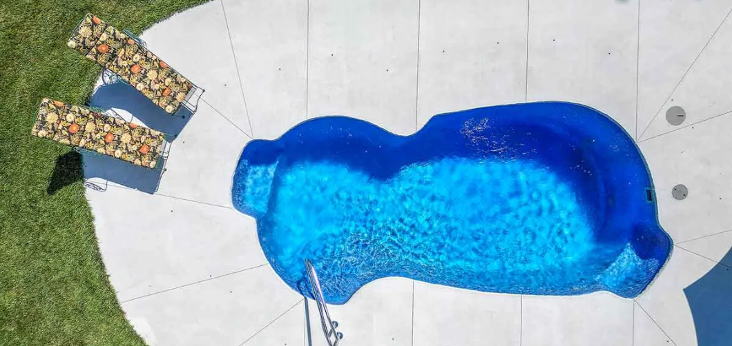 Overhead view of a blue, kidney-shaped swimming pool surrounded by white stone, with grassy area and steps.