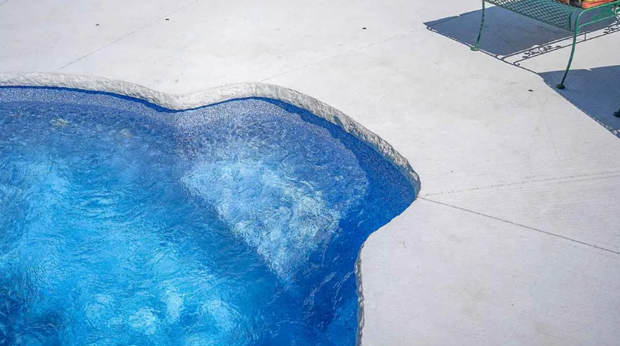 Blue swimming pool with a white concrete deck in bright sunlight.
